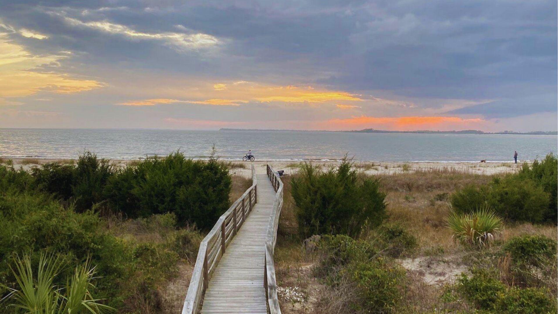 Wooden boardwalk leading through dunes to sandy beach at sunset with ocean and orange sky.