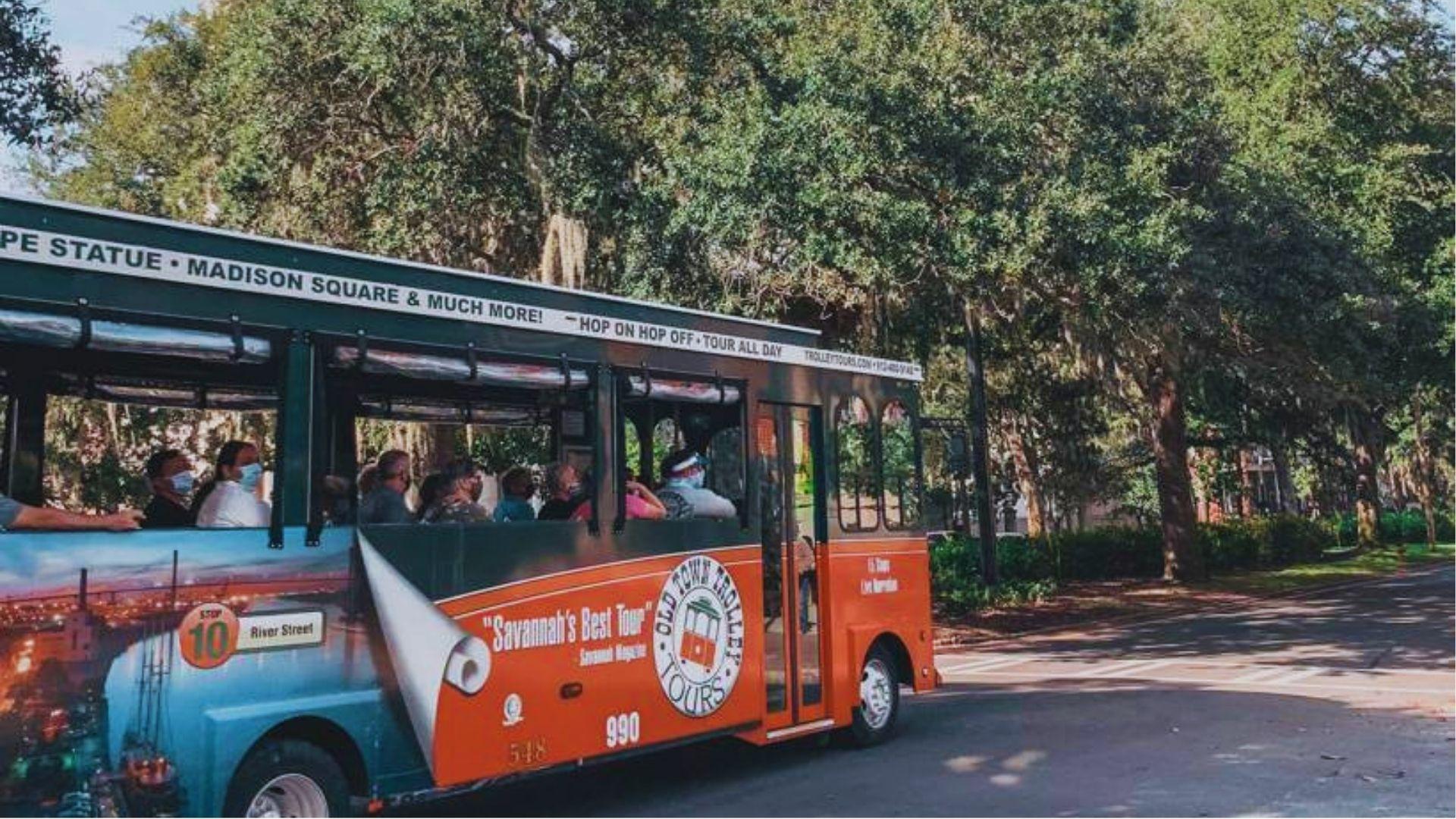 Orange Old Town Trolley tour bus carrying tourists along oak-lined historic Savannah street.