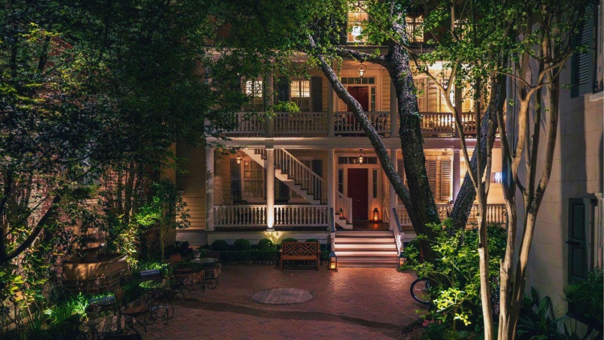 Historic two-story house illuminated at night with brick courtyard surrounded by trees and warm lighting.