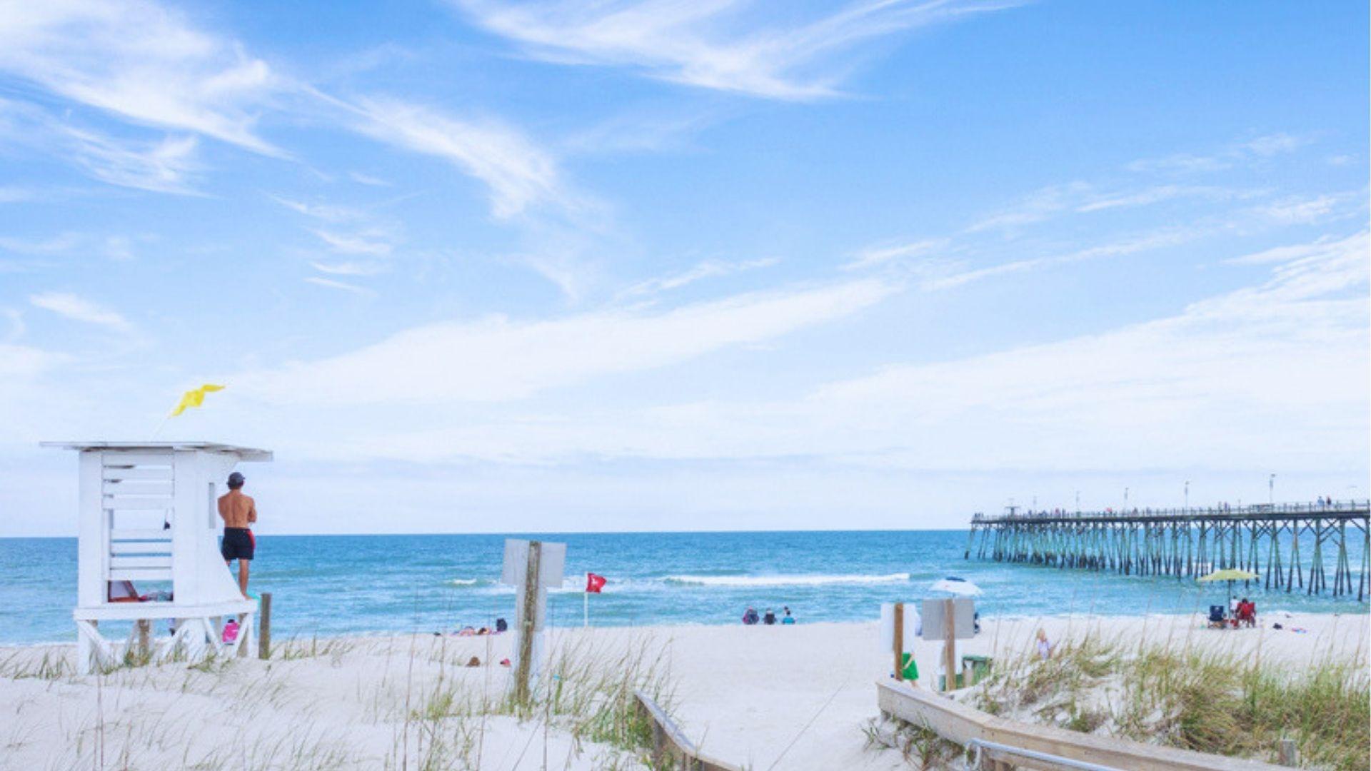 White lifeguard stand on sandy beach with yellow flag people relaxing and long fishing pier.