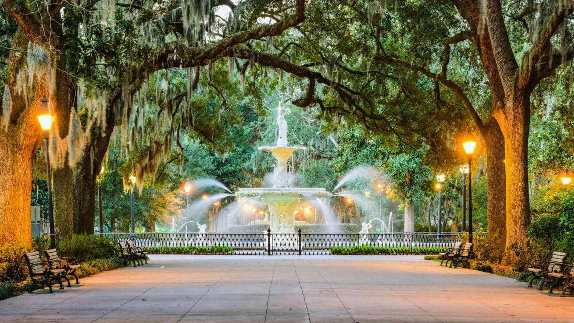 Lit fountain at dusk framed by mossy oaks, glowing lamps, benches and wide walkway