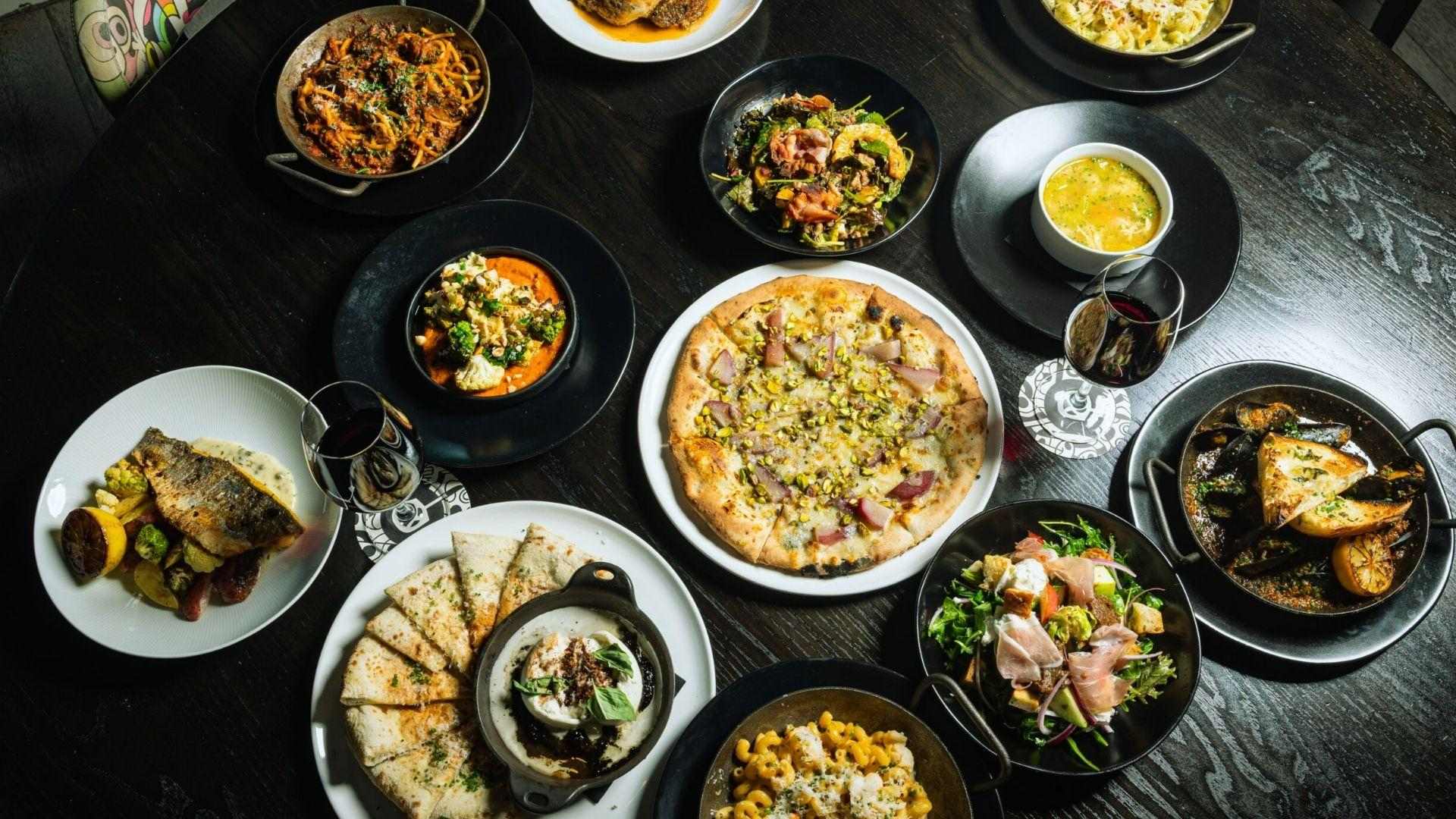 Overhead spread of pizza, pasta, fish and wine on dark table