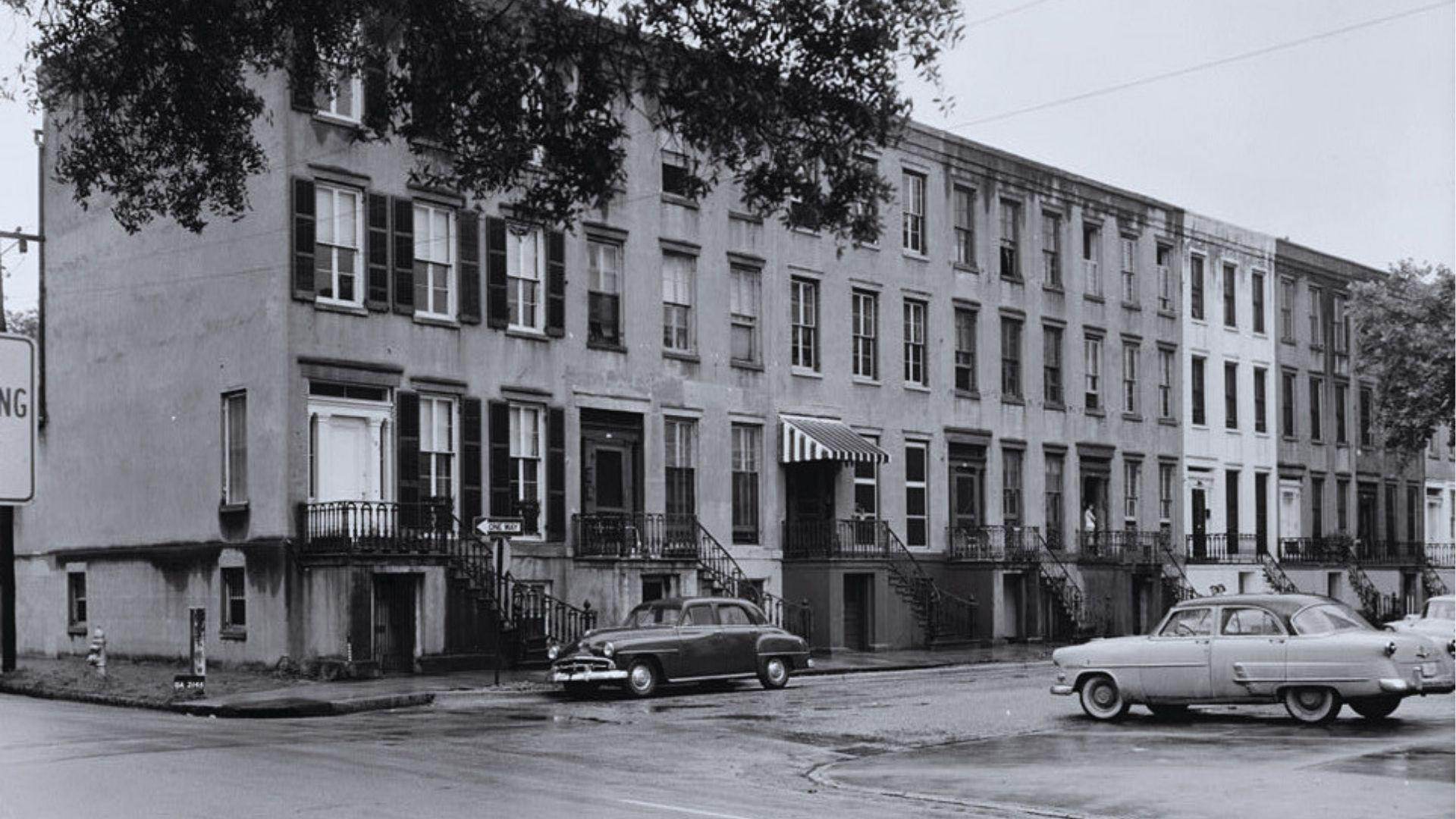 Black-and-white photo of historic row houses with vintage cars parked along wet city street today.