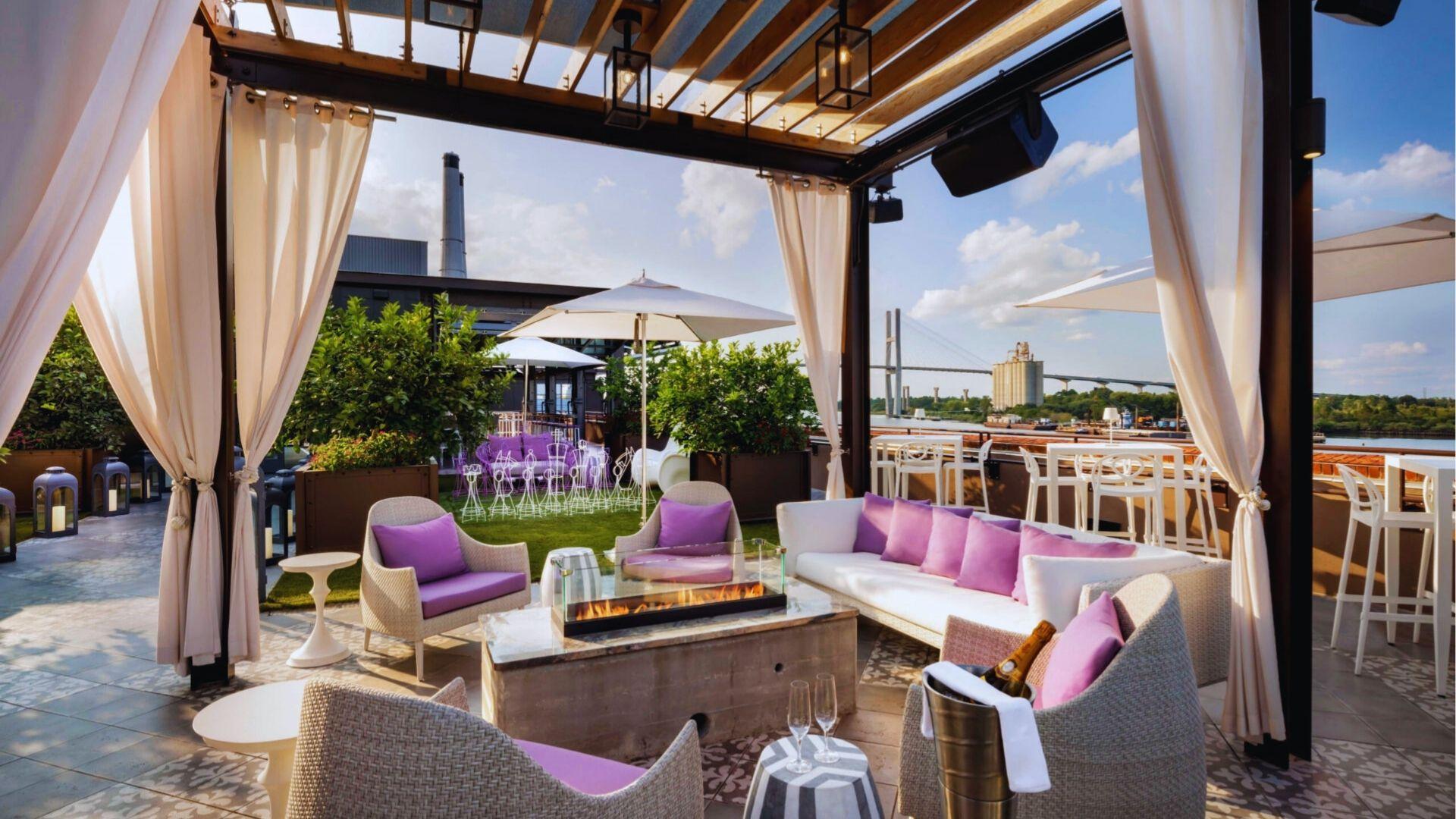 Rooftop lounge with purple cushions, fireplace, white curtains overlooking river and bridge at sunset today.