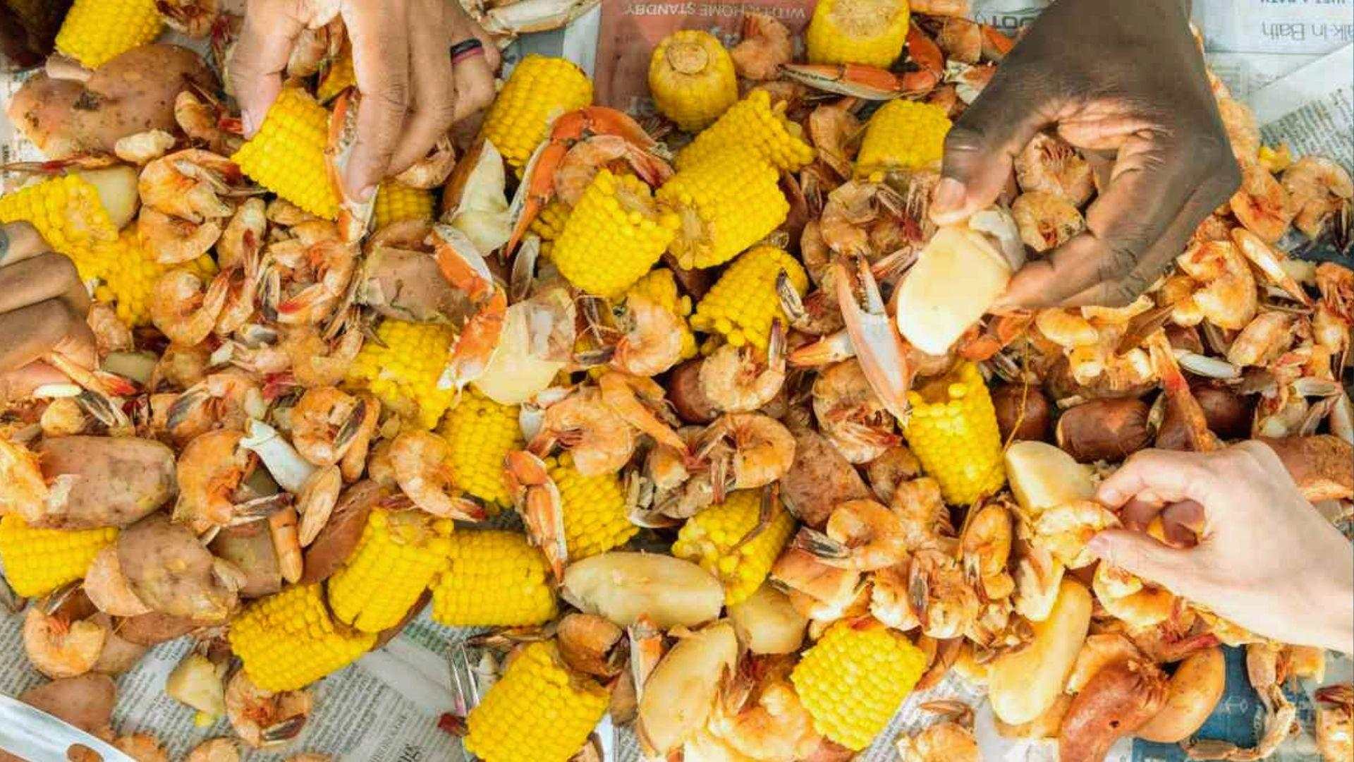 Hands reaching into Lowcountry boil spread of shrimp, corn, crab and potatoes on newspaper