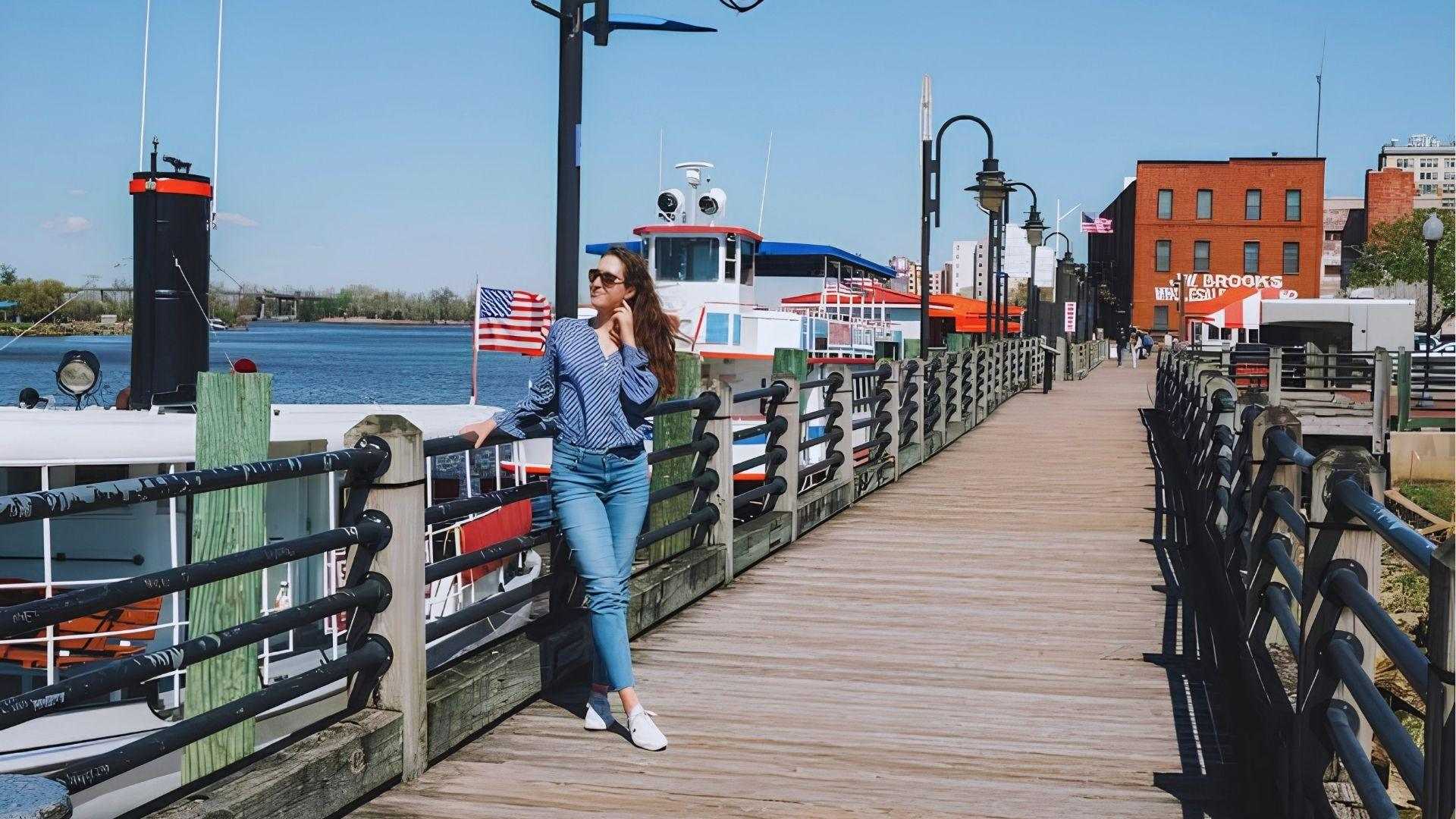 Woman in blue striped shirt posing on wooden boardwalk beside riverboats with American flag waving.