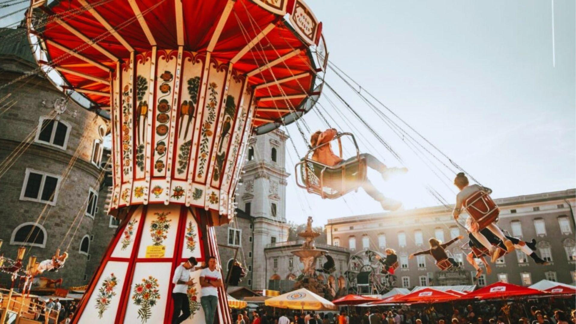 Riding ornate red and white swing carousel at sunny European-style festival fairground with historic buildings.