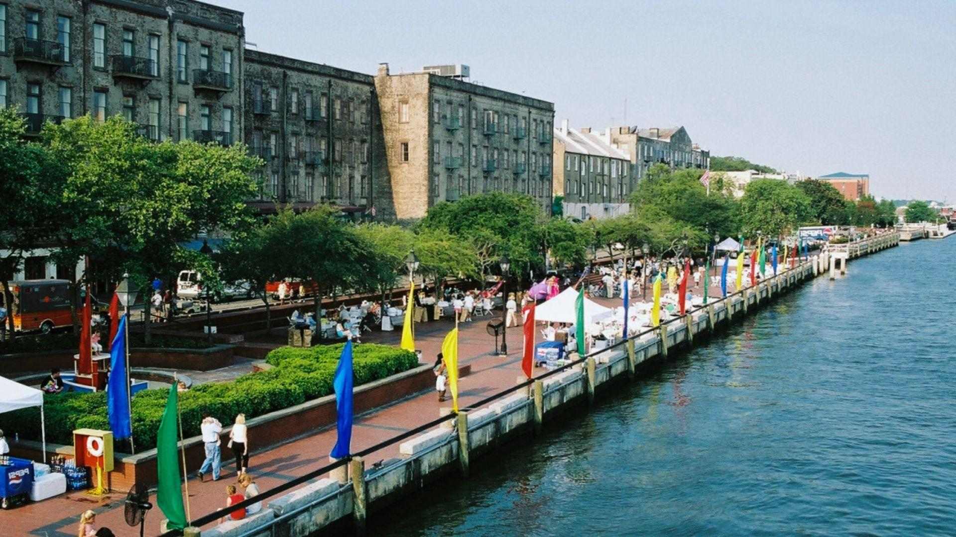 Riverfront promenade with colorful flags, crowds, historic brick warehouses along water at daytime festival