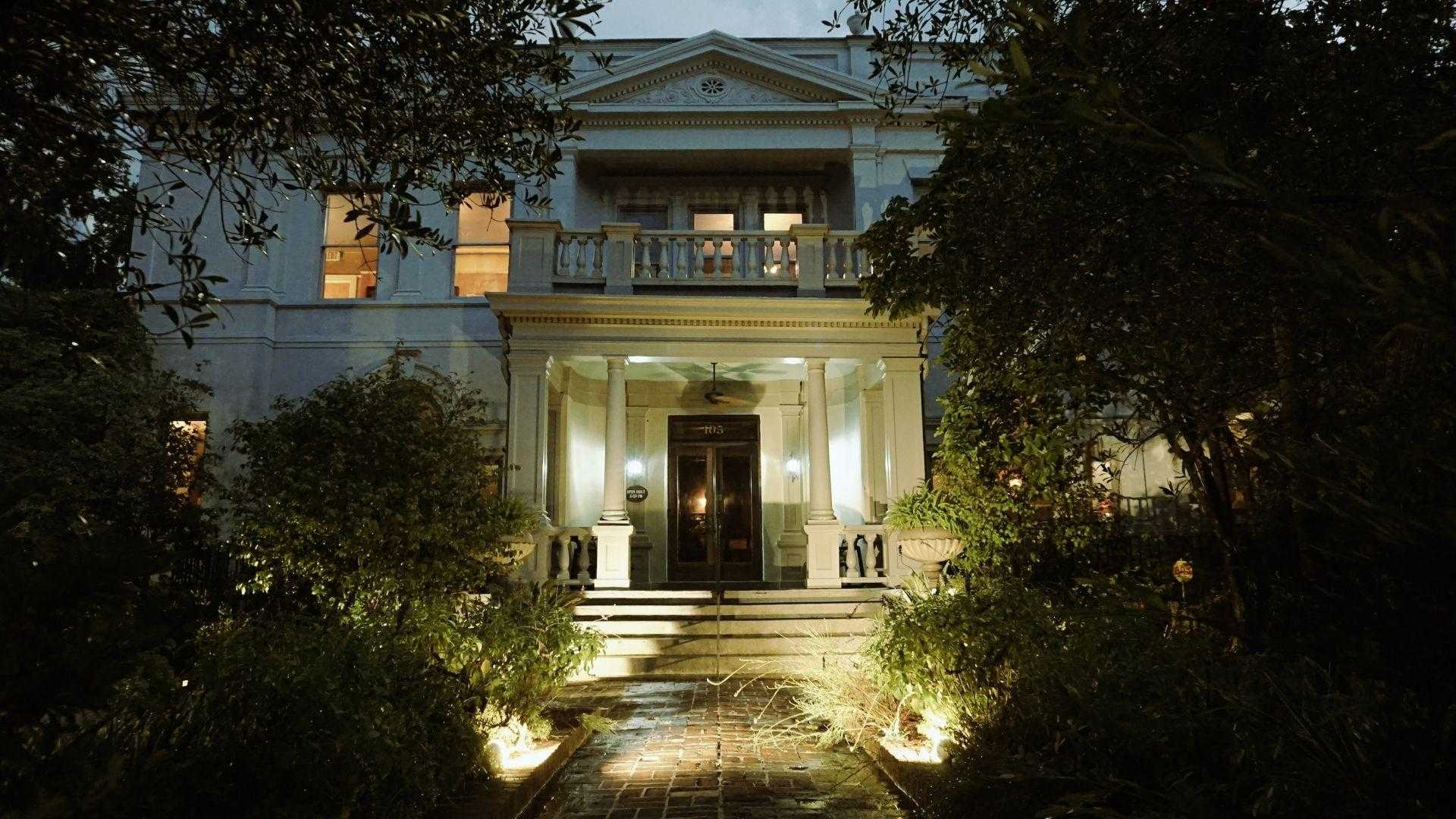 Stately white mansion at dusk with illuminated columned portico, glowing windows, brick pathway lined with garden lights and lush trees.