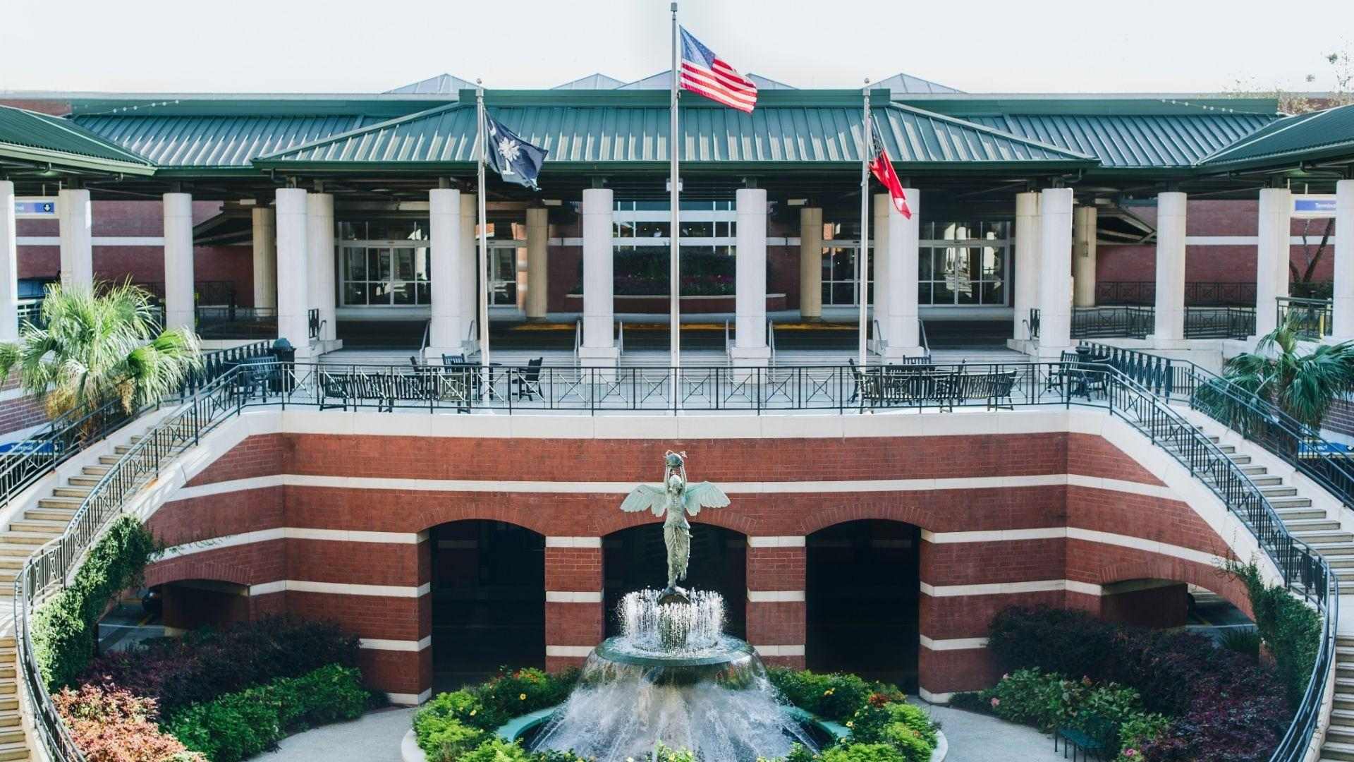 Brick plaza with twin curved stairs, central angel fountain, flags above colonnaded building and greenery