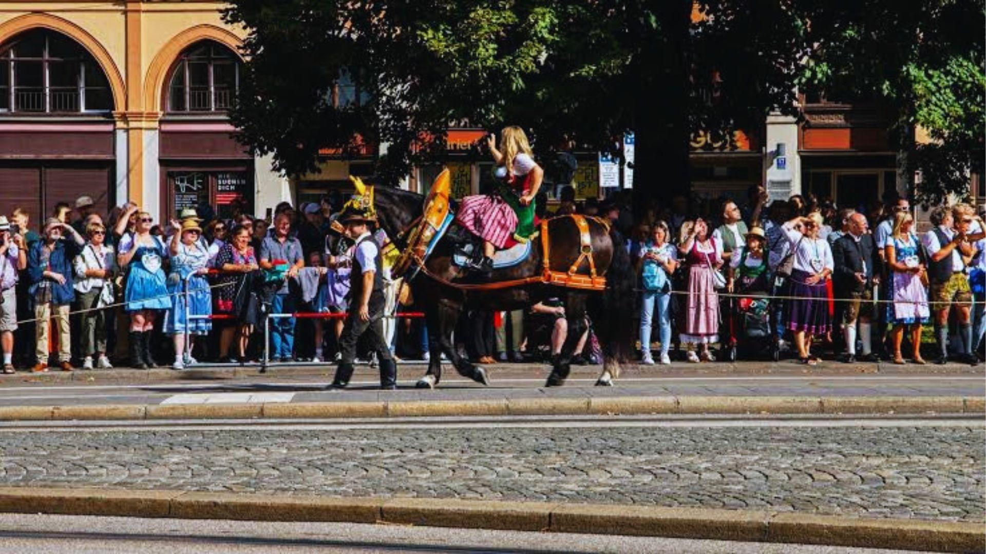 Woman riding decorated black horse in parade while crowd watches from sidewalk on sunny day.
