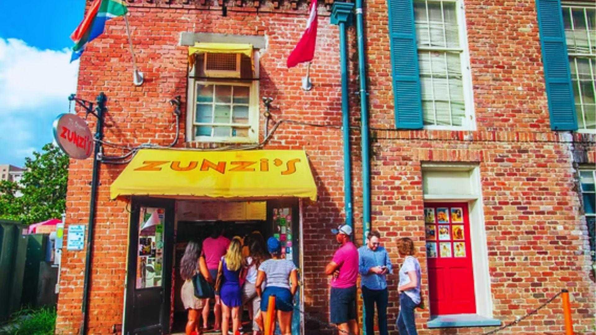 Crowd waiting outside Zunzi's restaurant under bright yellow awning on sunny red brick building today.