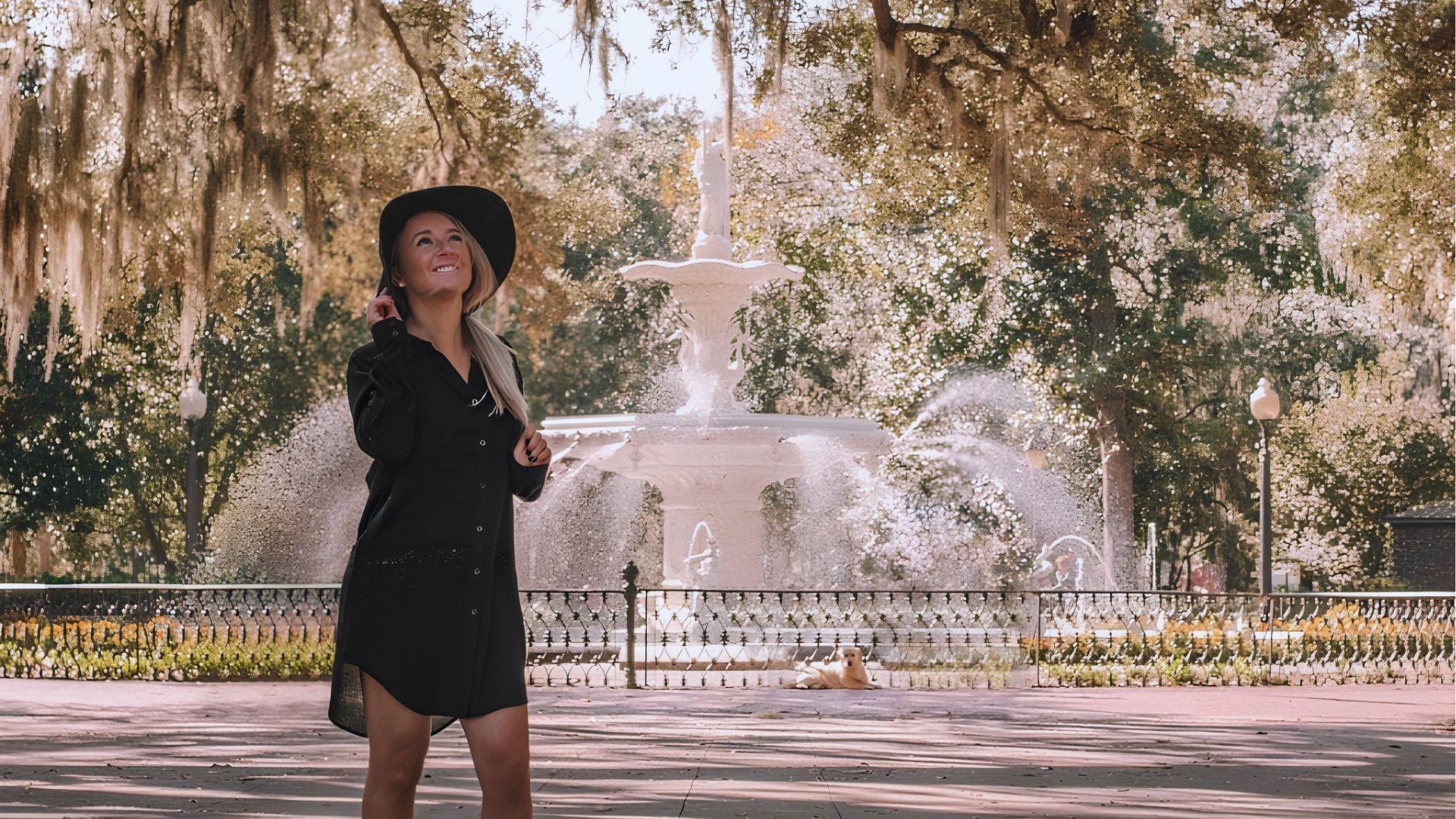 Smiling woman in black dress and hat posing before Forsyth Park fountain surrounded by trees.
