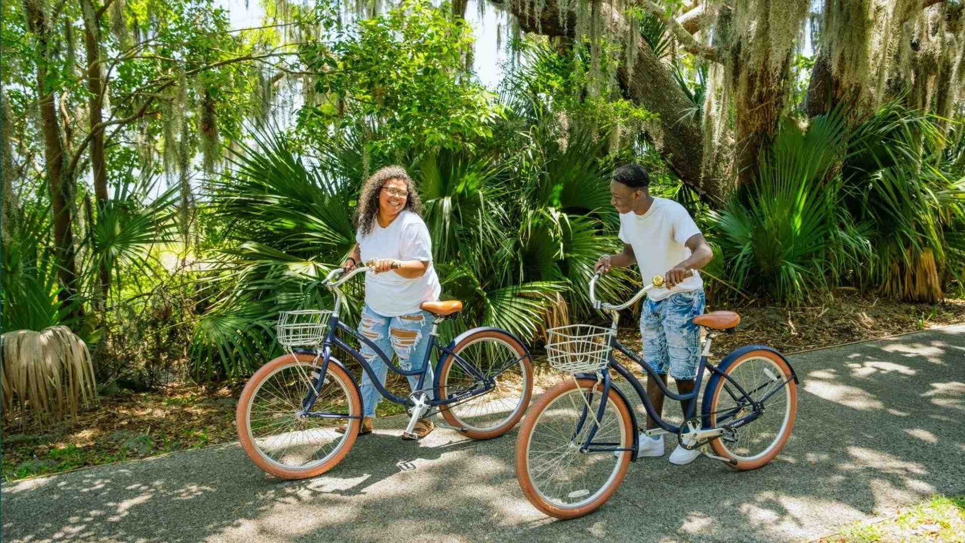 Two smiling people with blue beach cruisers on shaded path under mossy oaks and palms
