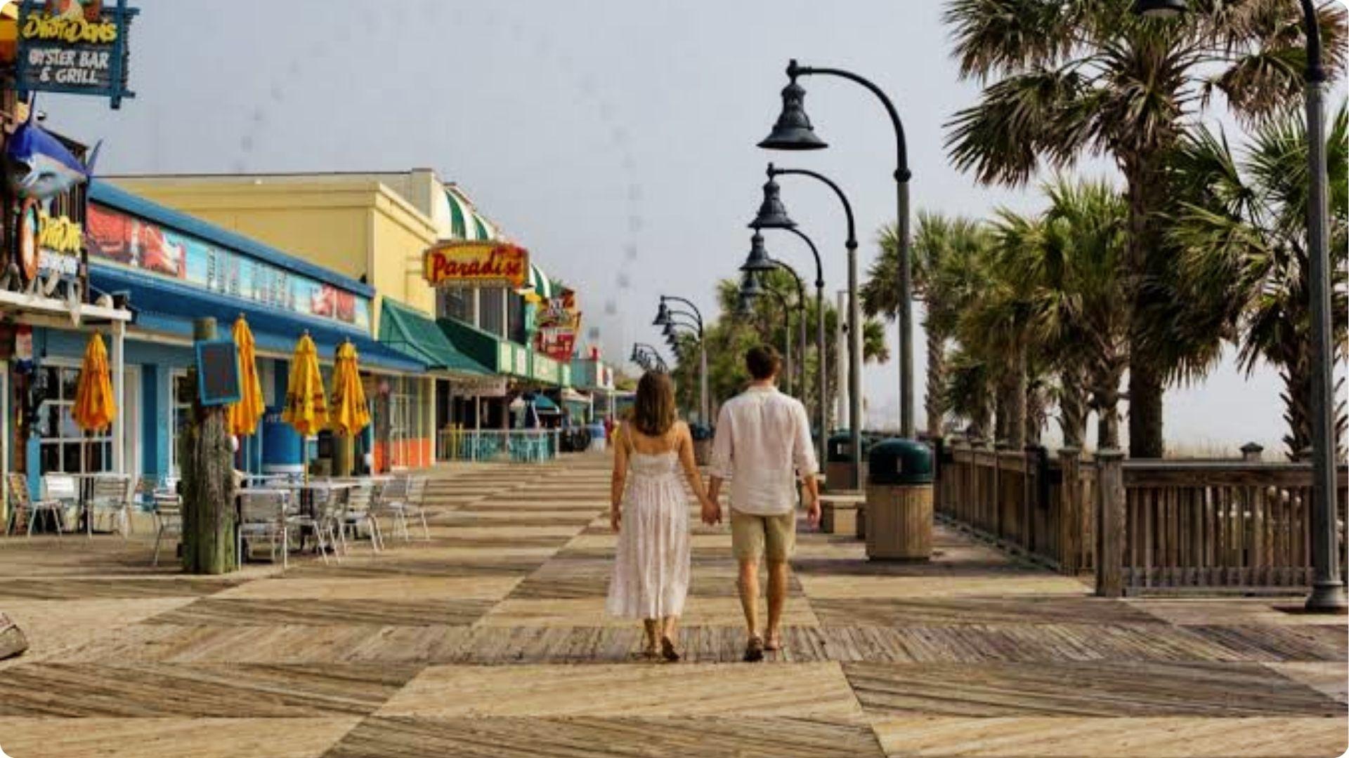 Couple holding hands walking along Myrtle Beach boardwalk lined with palm trees and seaside shops