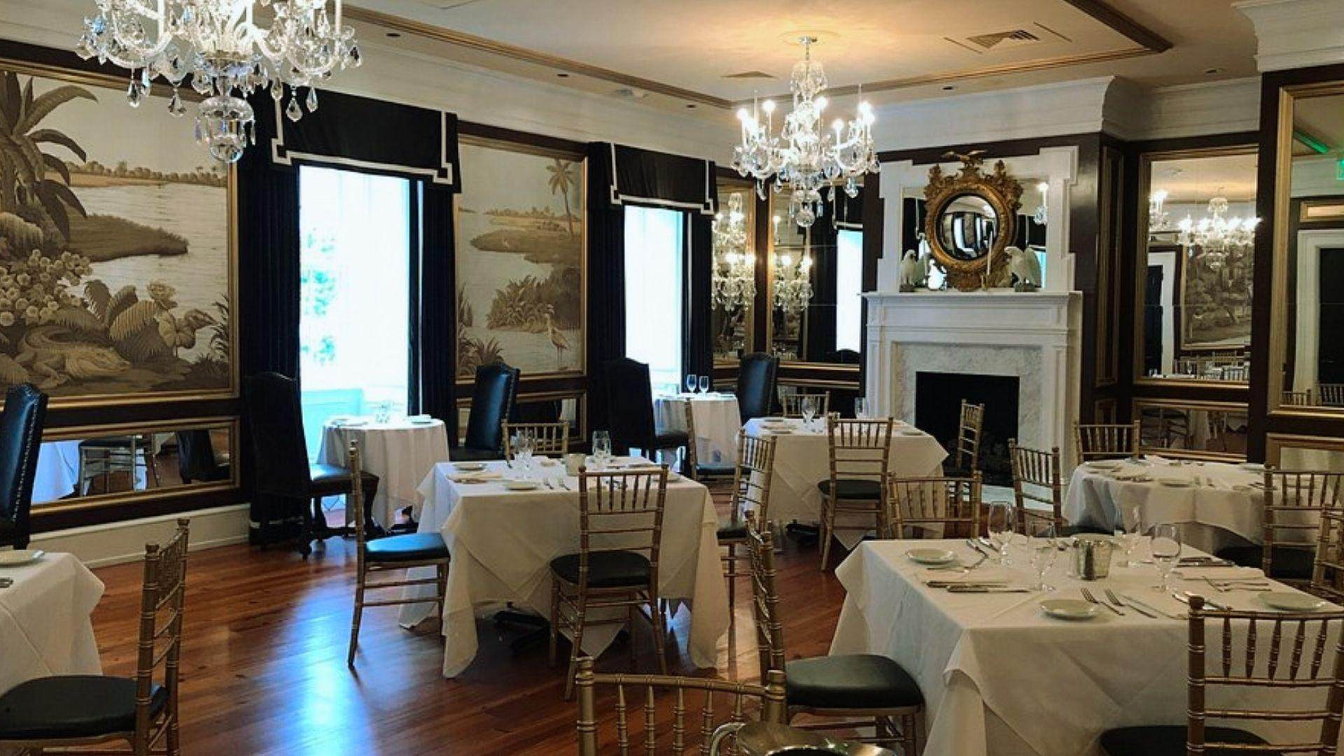 Elegant formal dining room with crystal chandeliers, white-linen tables, gold chiavari chairs, black valances, scenic mural wallpaper.