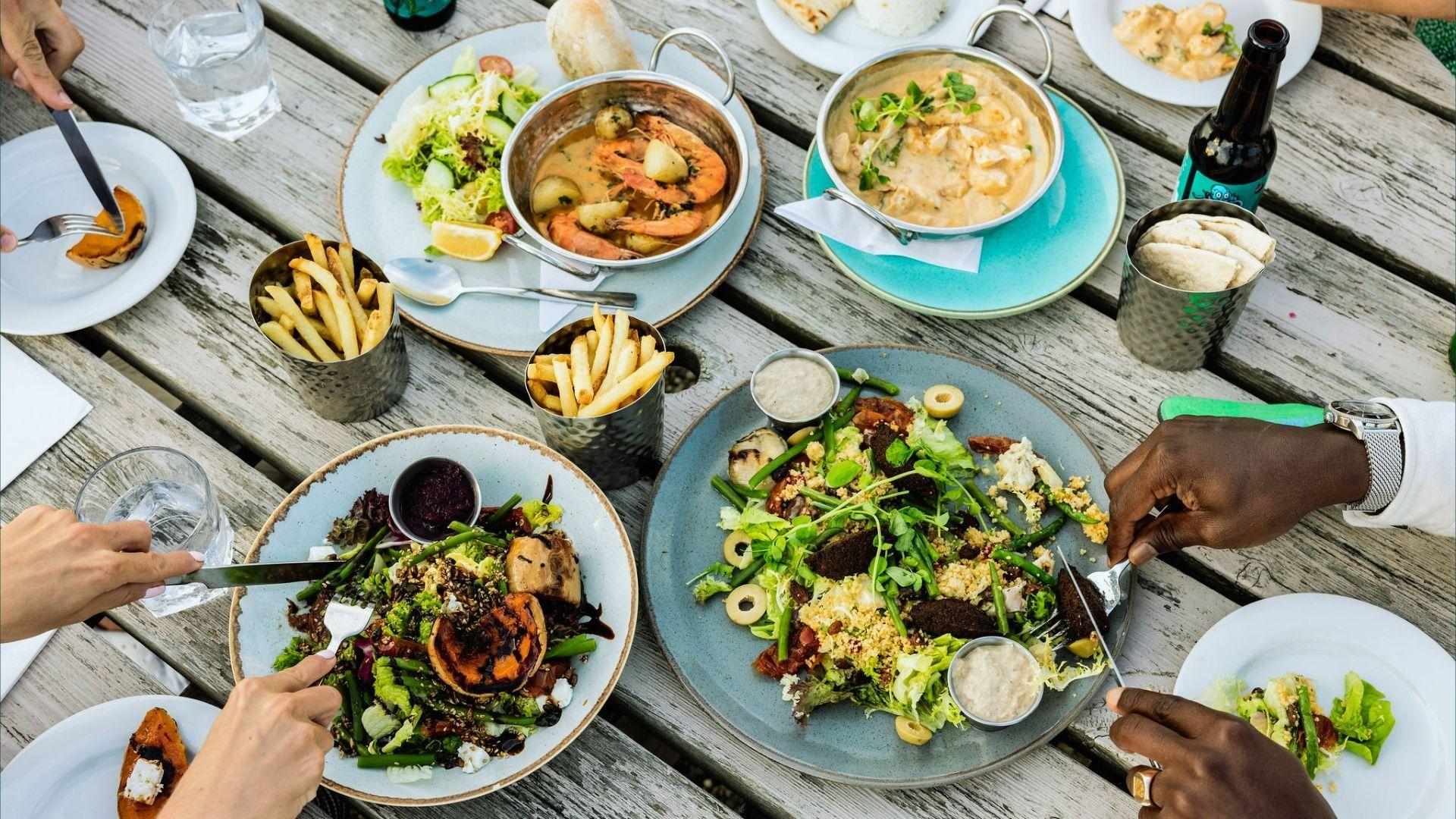 Overhead group lunch with salads, shrimp curry, fries, pita and beer on rustic table