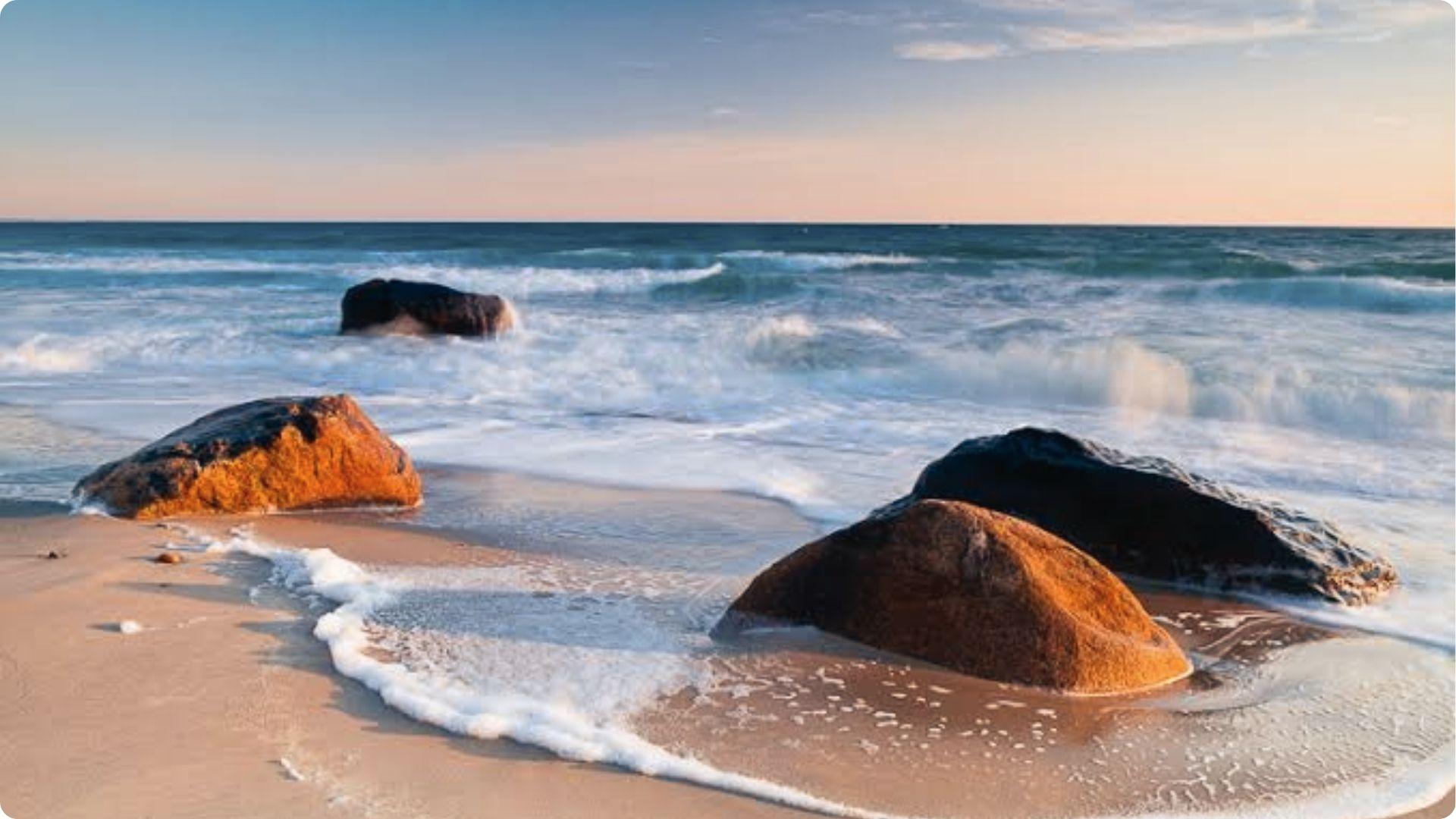 Waves crashing on sunlit rocks at East Beach St Simons shoreline with gentle sea foam at dawn.