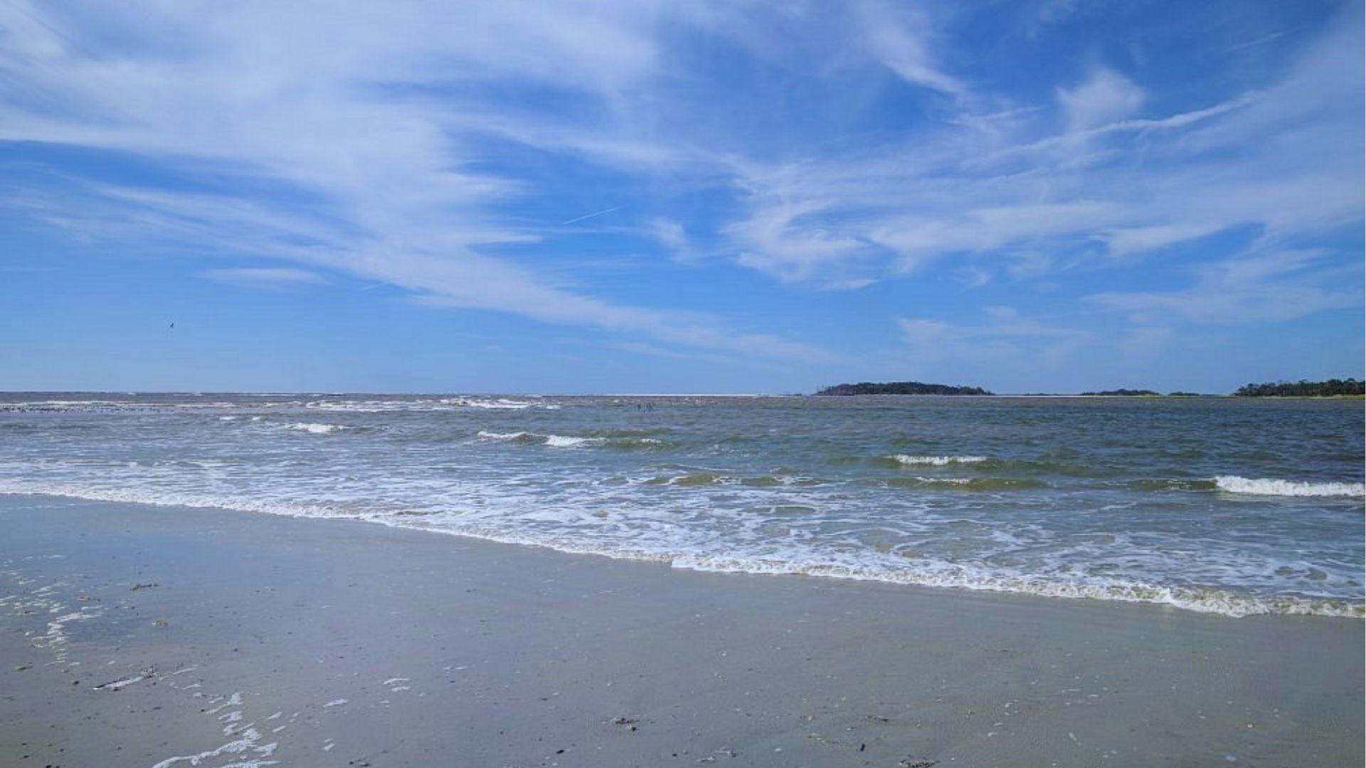 beach with gentle waves lapping gray sand under wispy blue sky and distant islands.