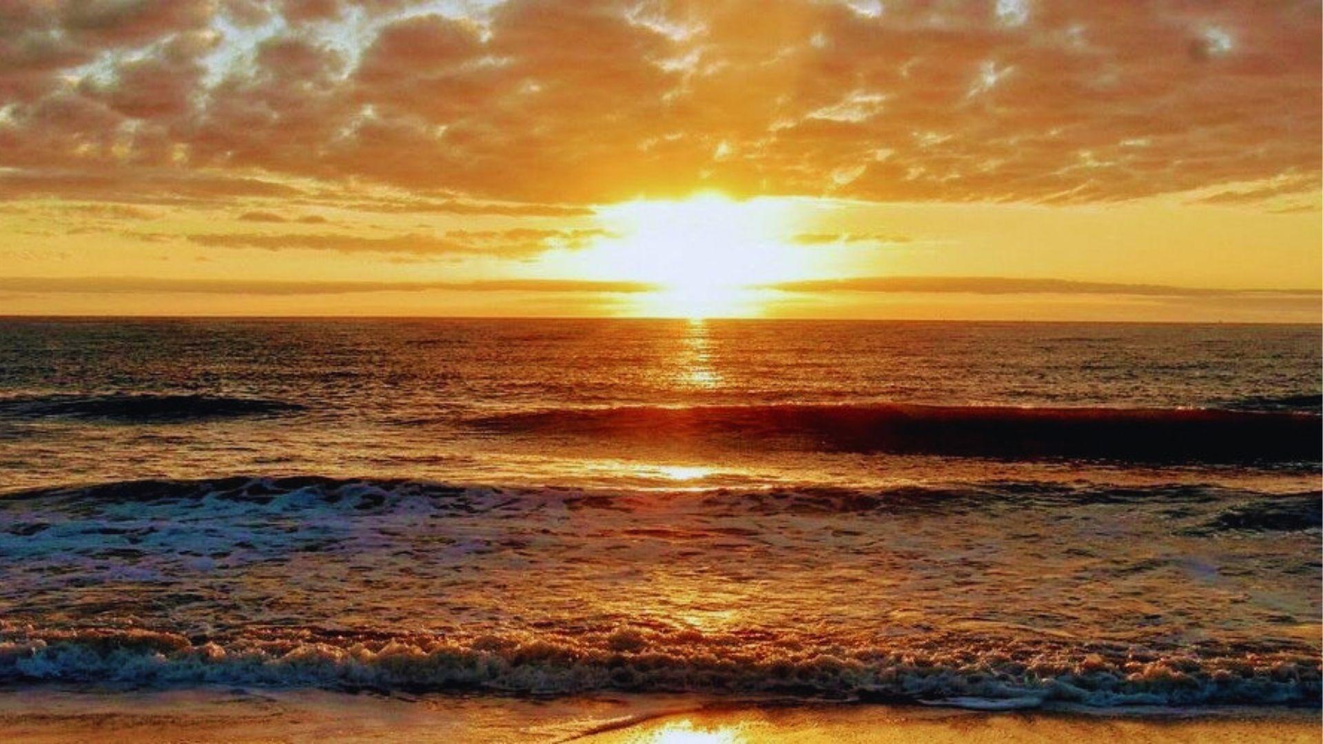 Sunset over ocean waves with glowing sky reflecting on wet sandy beach and foamy surf.