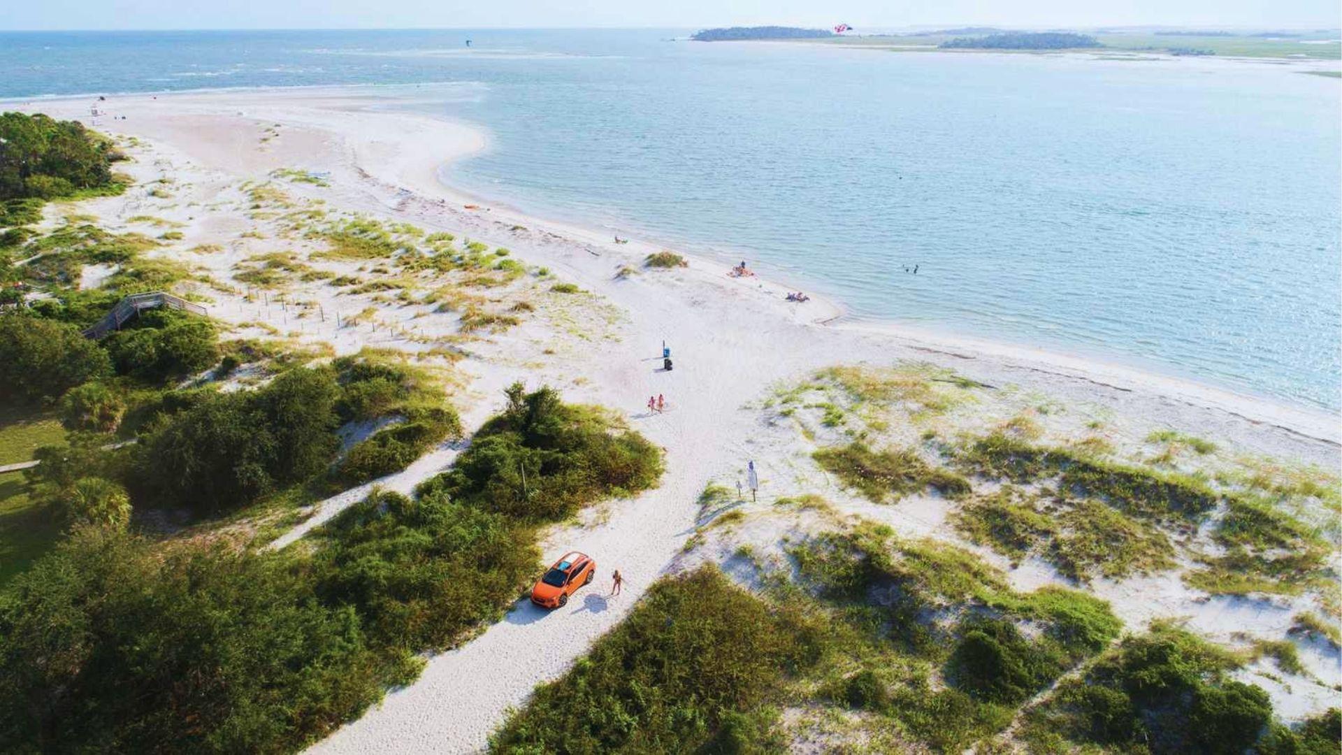 View of white sandy beach peninsula with dunes, greenery, orange car and calm blue ocean.