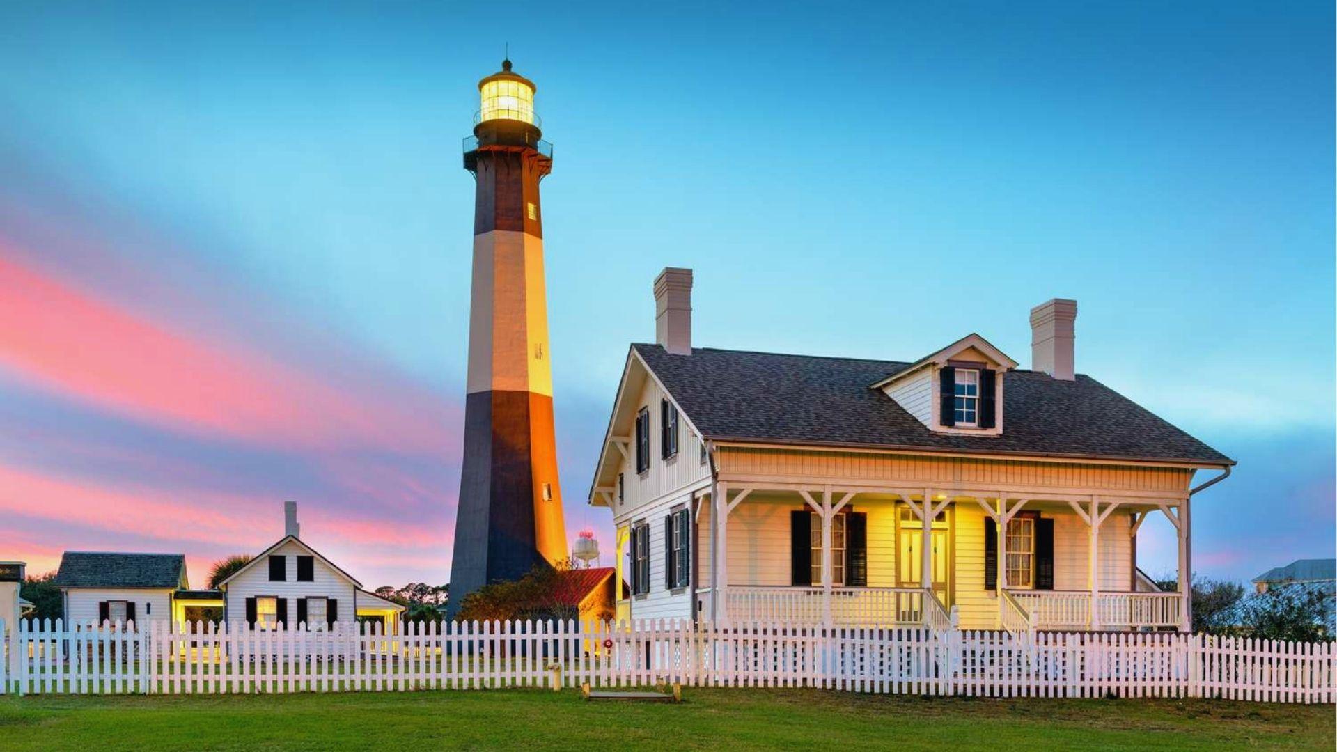 Historic Tybee Island Lighthouse glowing at dusk beside keeper's house with pink sunset sky behind.