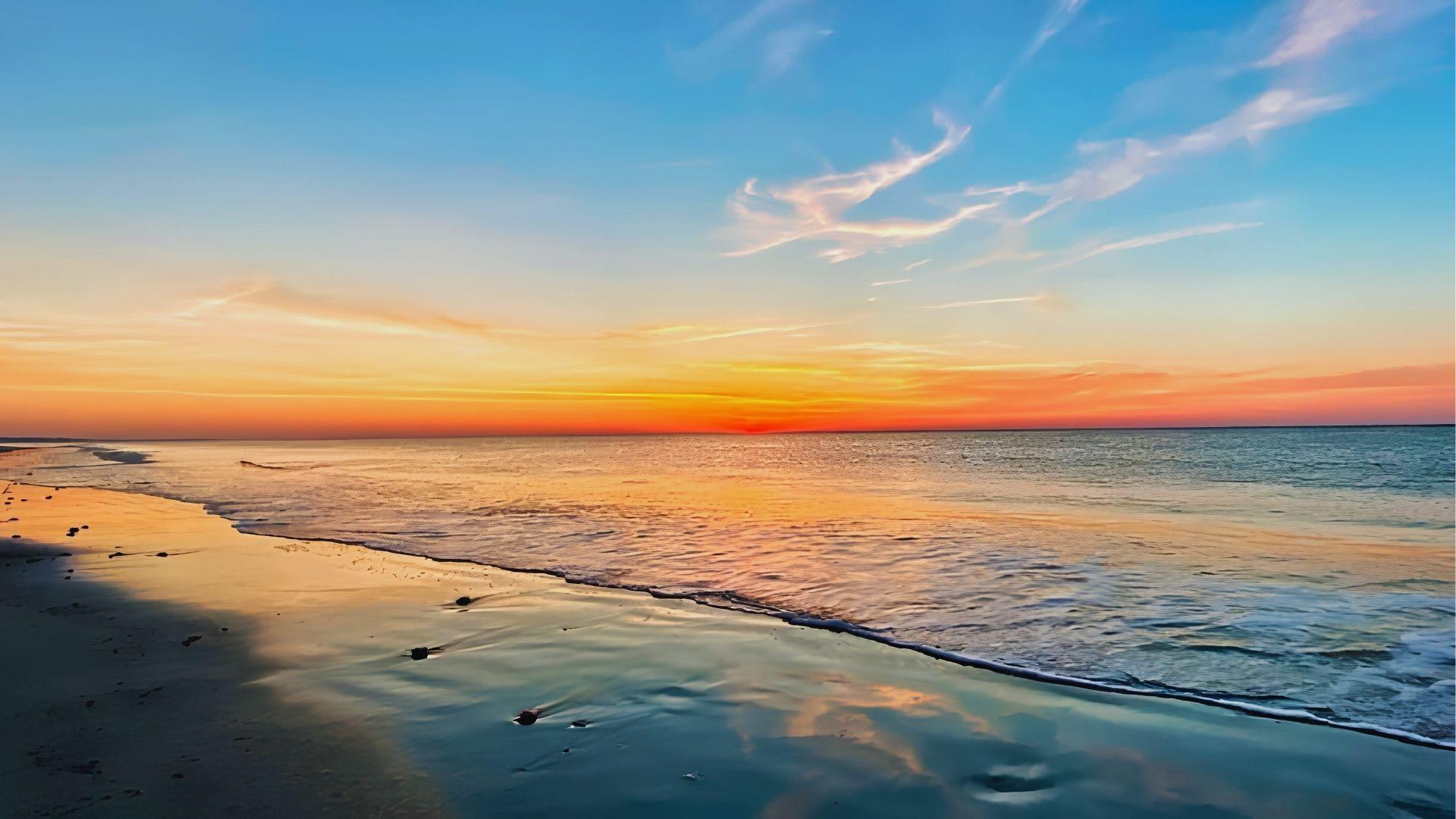 Sunset over ocean with orange pink sky reflecting on wet sandy shoreline and gentle waves.