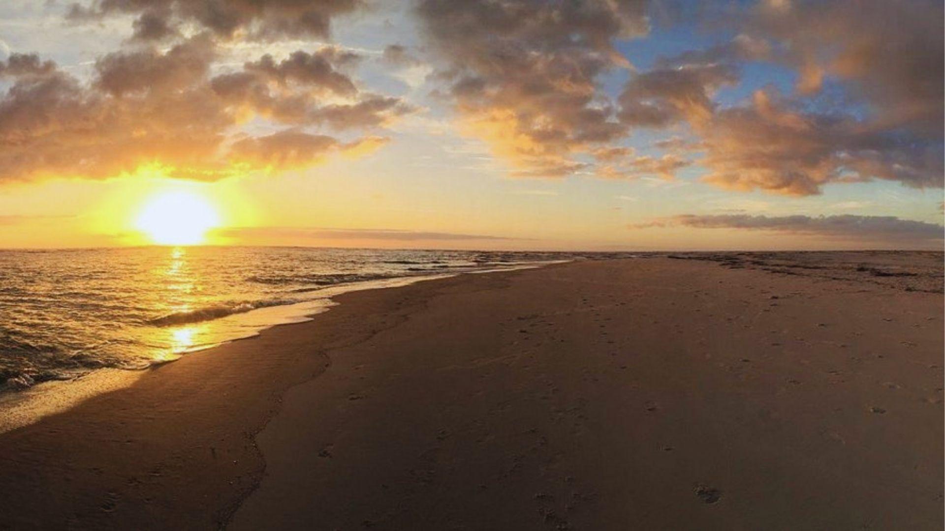 Golden sunrise over calm ocean waves lapping empty sandy beach with footprints stretching into distance.