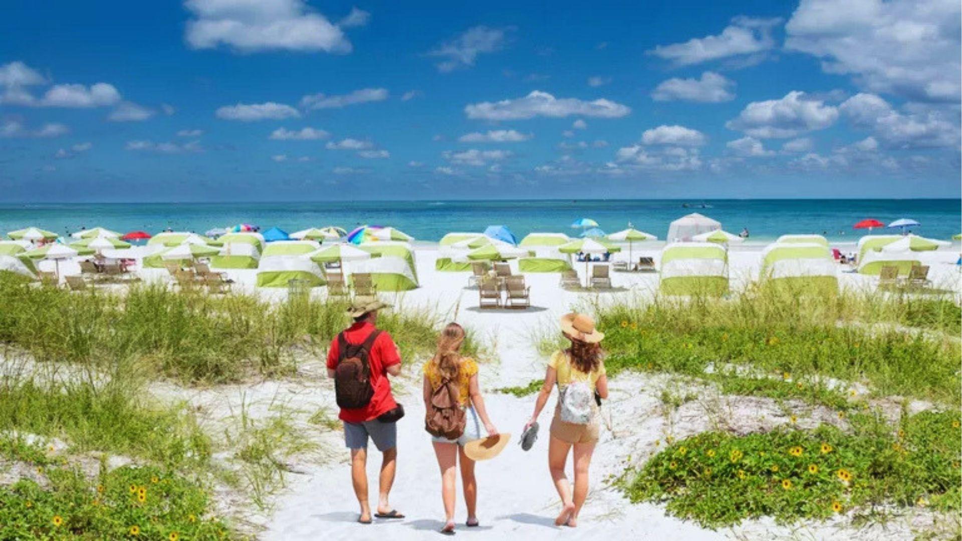 Travelers walking down a sandy path toward white beach lined with green-yellow cabanas and turquoise ocean.