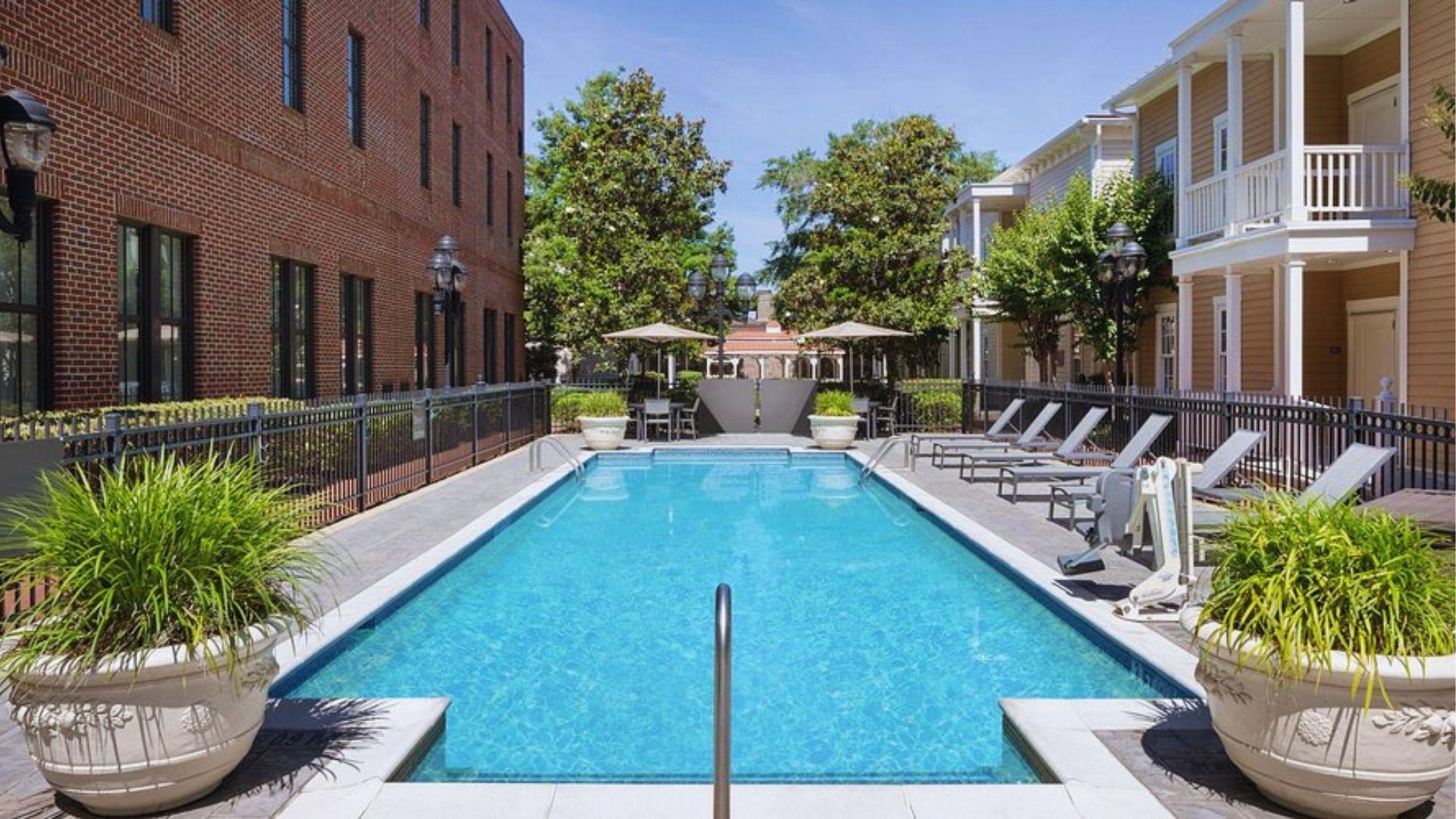 Swimming pool with clear blue water lounge chairs potted plants between brick and beige buildings.