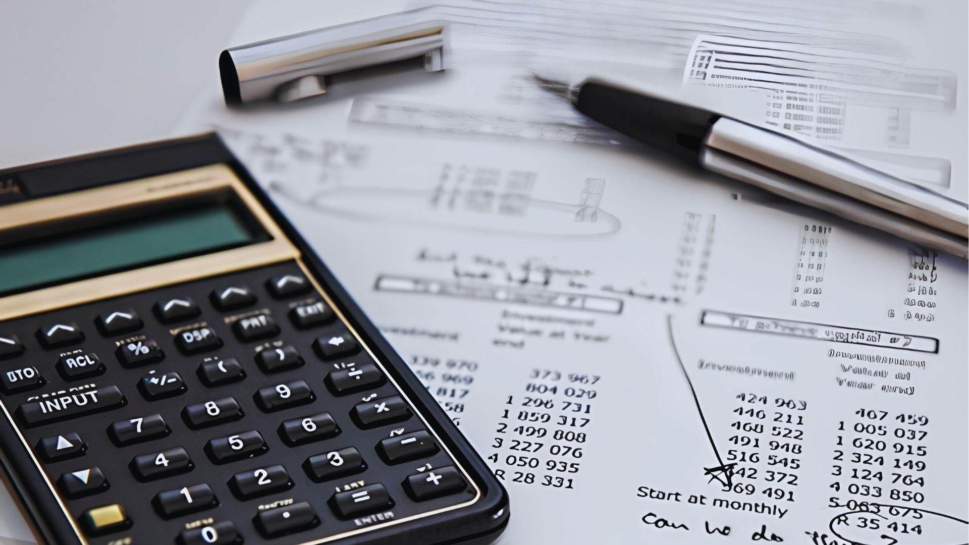 Calculator resting on investment documents with handwritten notes, pens and numerical charts for tax calculations.