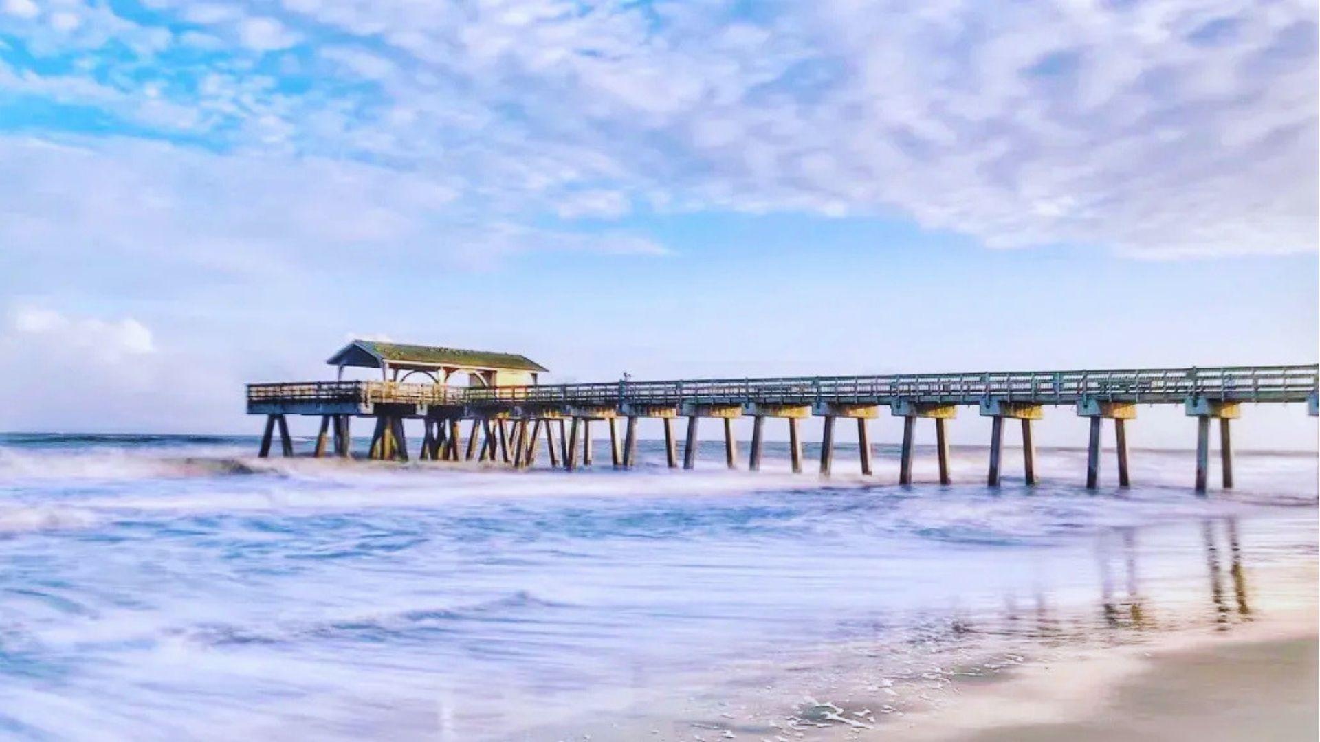 Wooden pier extending over foamy ocean waves under bright blue sky with scattered white clouds.