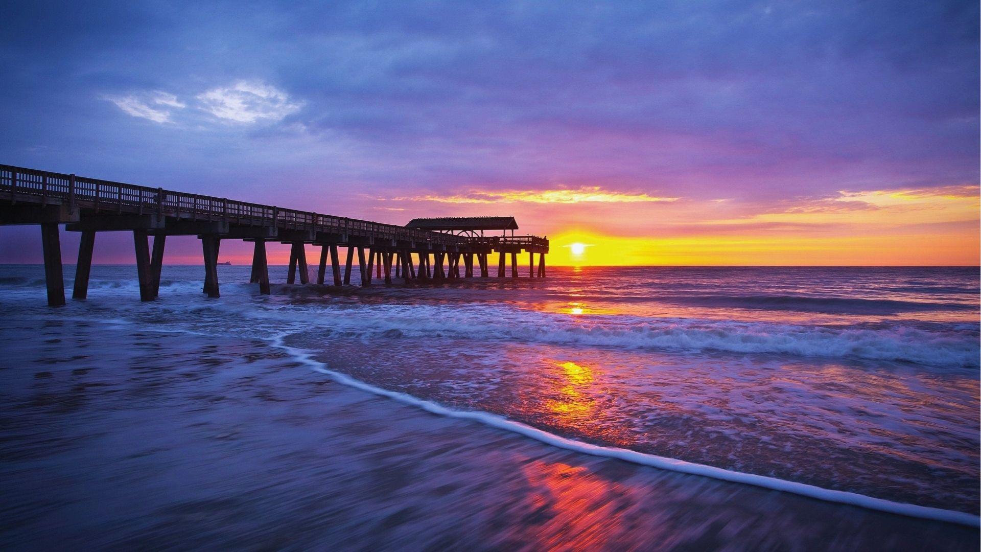 Wooden pier extending over ocean waves at vibrant orange purple sunset reflecting on wet sand.