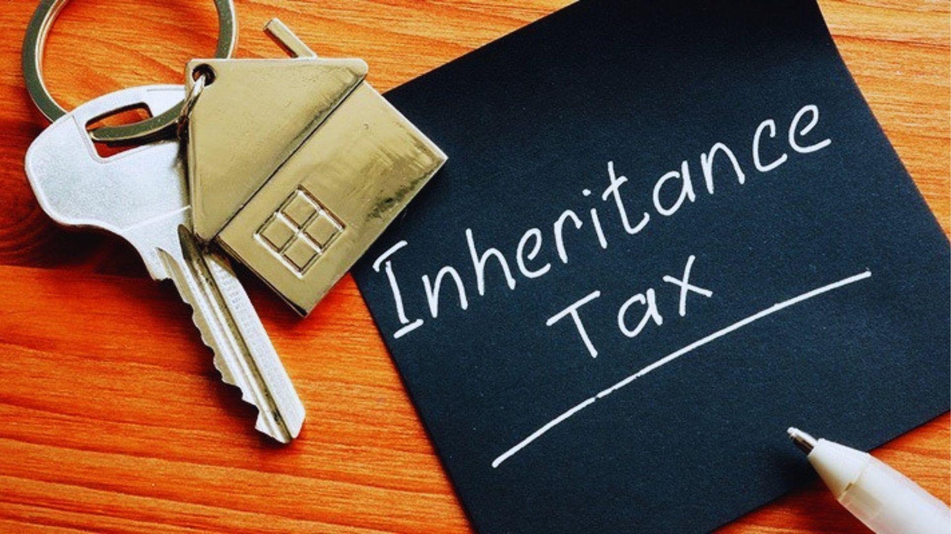 House-shaped keychain on keys next to black card handwritten Inheritance Tax and pen on wood.