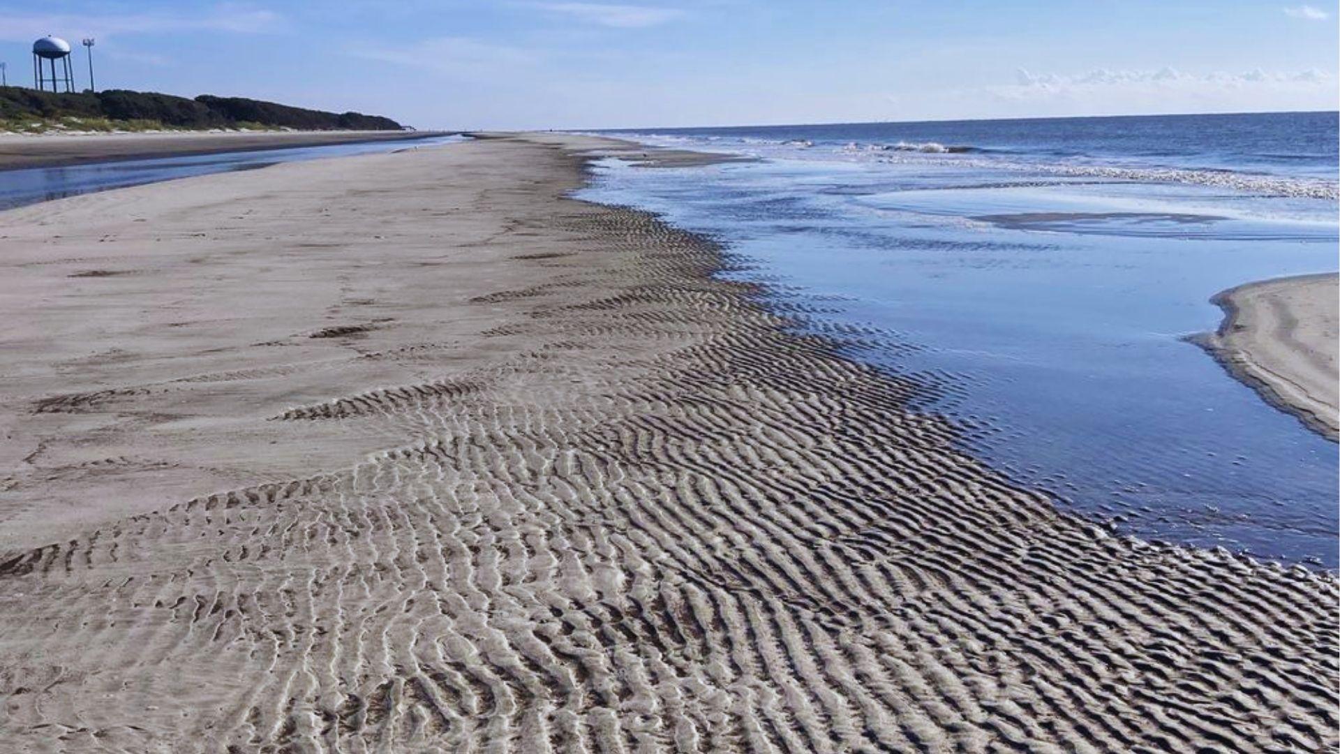 Sandbar stretching along beach with shallow water pools and distant water tower under blue sky.