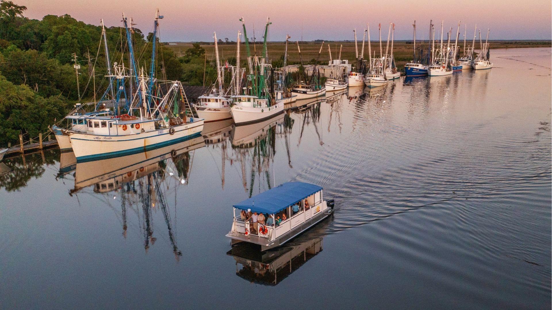 Shrimp boats moored along marshy riverbank with tour boats cruising past at glowing sunset reflection.