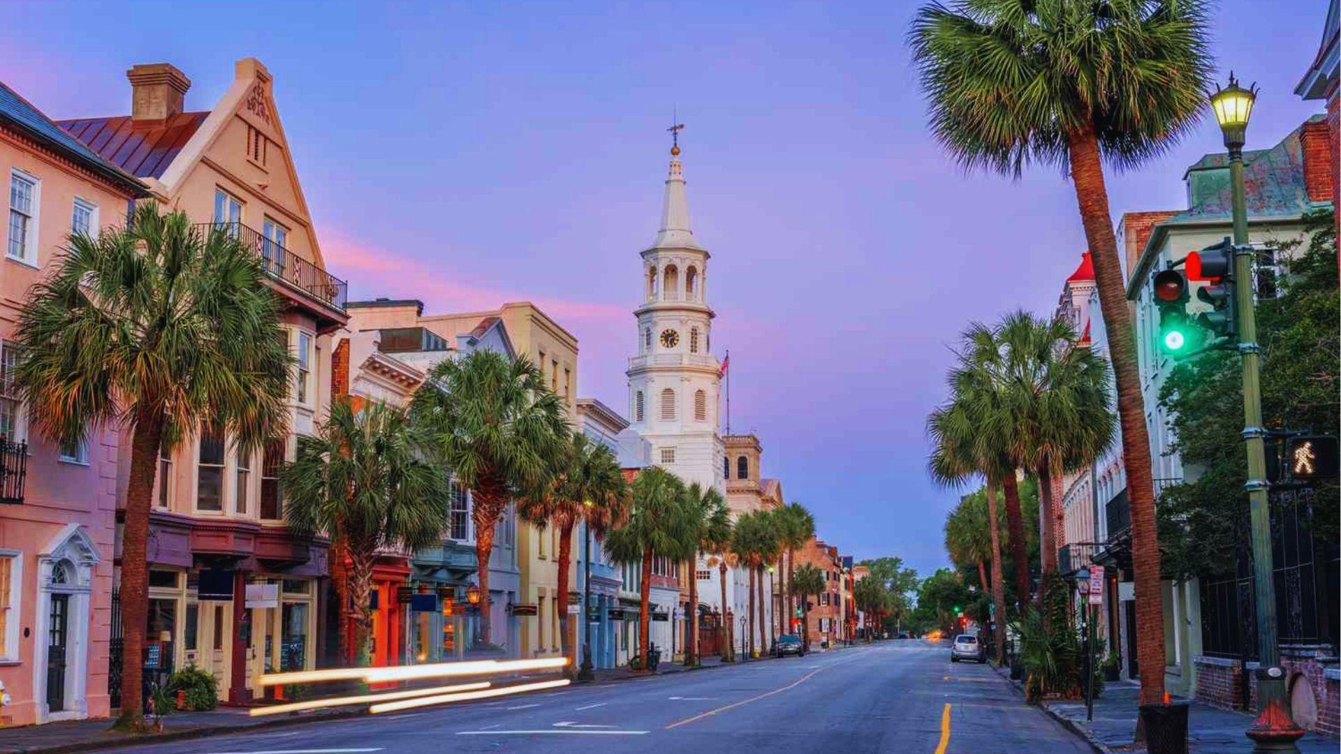 Charleston street at twilight with pastel buildings palm trees and white church steeple in background.
