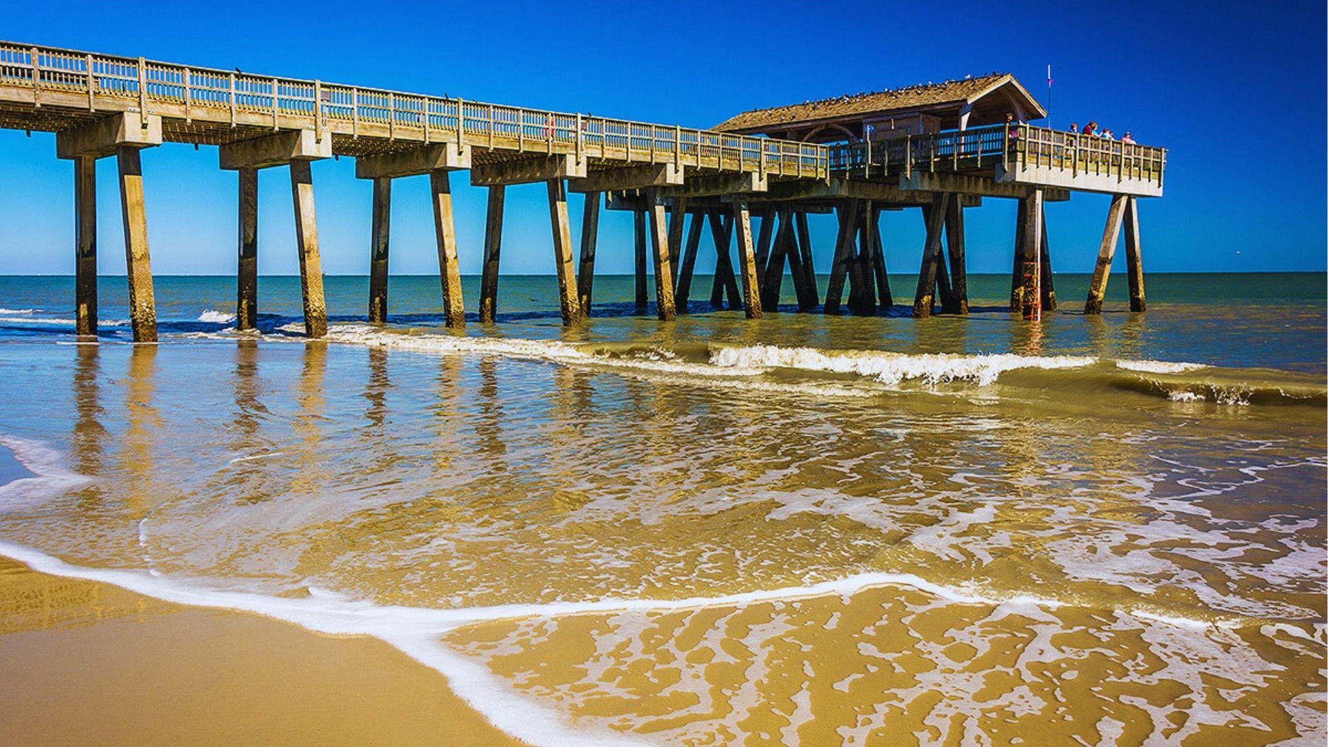 Pier with shelter standing over shallow surf on sandy beach under clear vibrant blue sky.