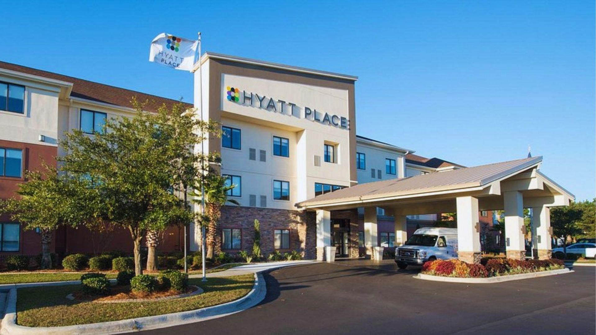 Hyatt Place hotel exterior with portico shuttle van palm trees and landscaping under sunny sky.