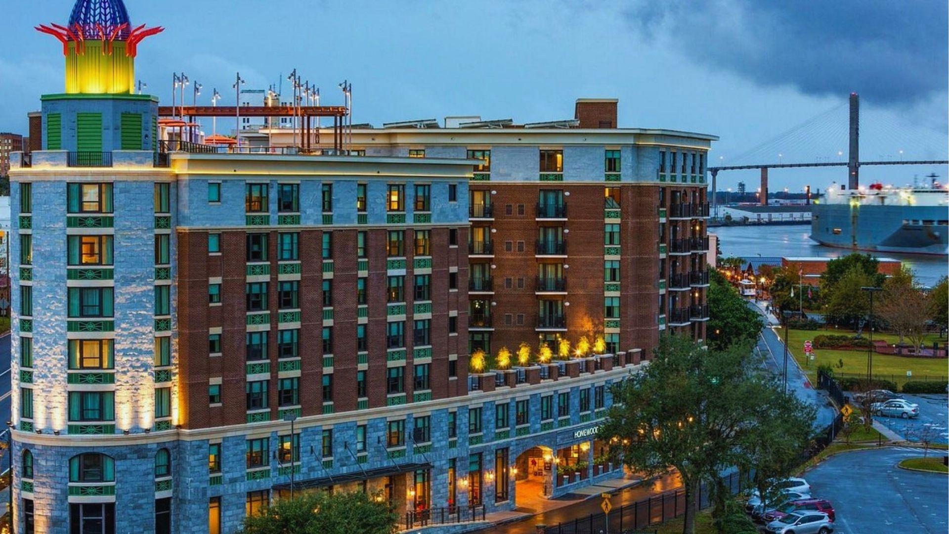 View of colorful Homewood Suites hotel with illuminated tower river bridge and cargo ship nearby.