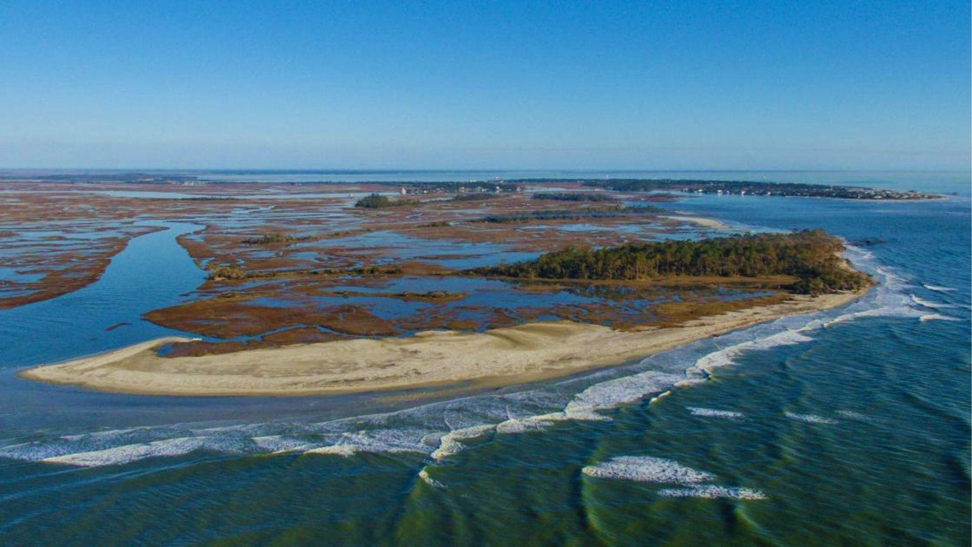 View of coastal barrier island with sandy beaches, marsh wetlands and ocean waves meeting shoreline.