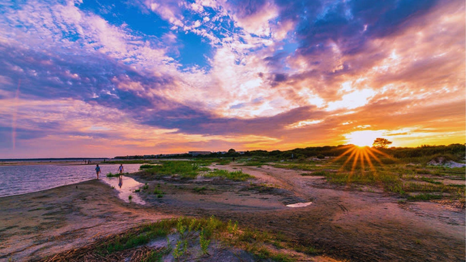 Sunset casting golden rays over the beach with two people wading through shallow tidal waters.