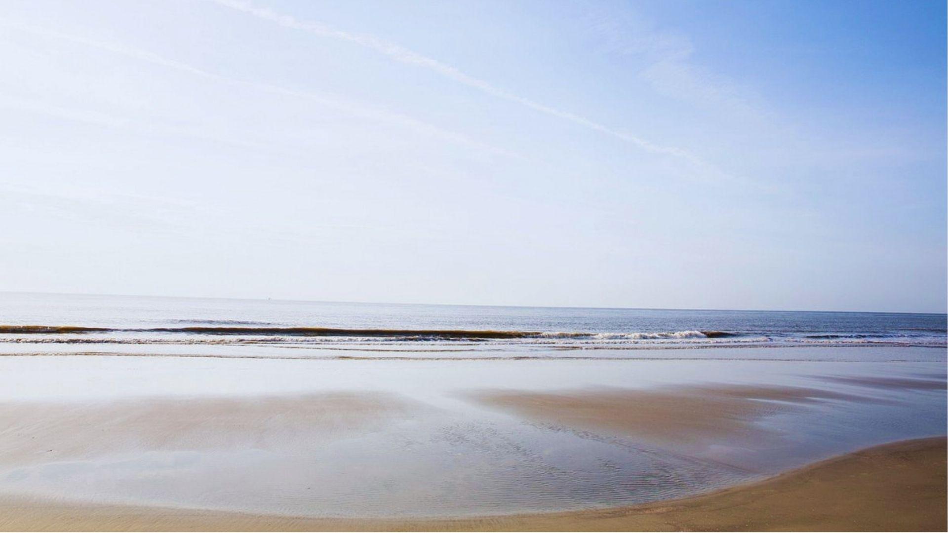 Wide empty beach with glassy tidal pools reflecting blue sky and gentle waves rolling offshore.
