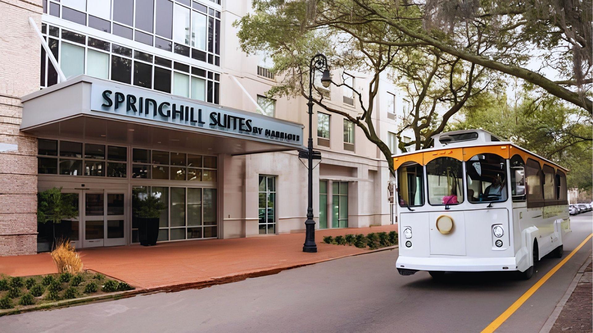 White trolley tour passing SpringHill Suites by Marriott entrance on tree-lined Savannah downtown sunny street.