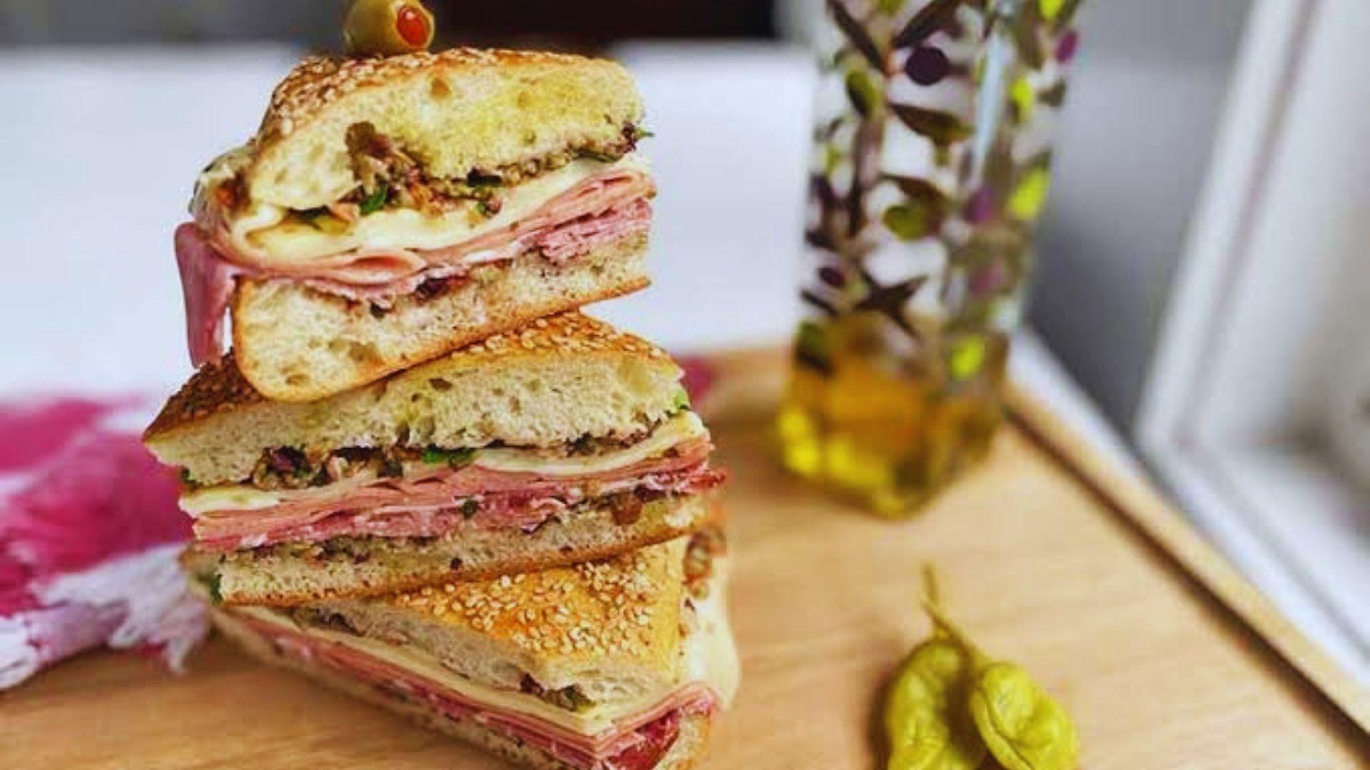 A round muffuletta sandwich cut in half, showing layers of deli meats, provolone, and olive salad on a sesame seed roll.