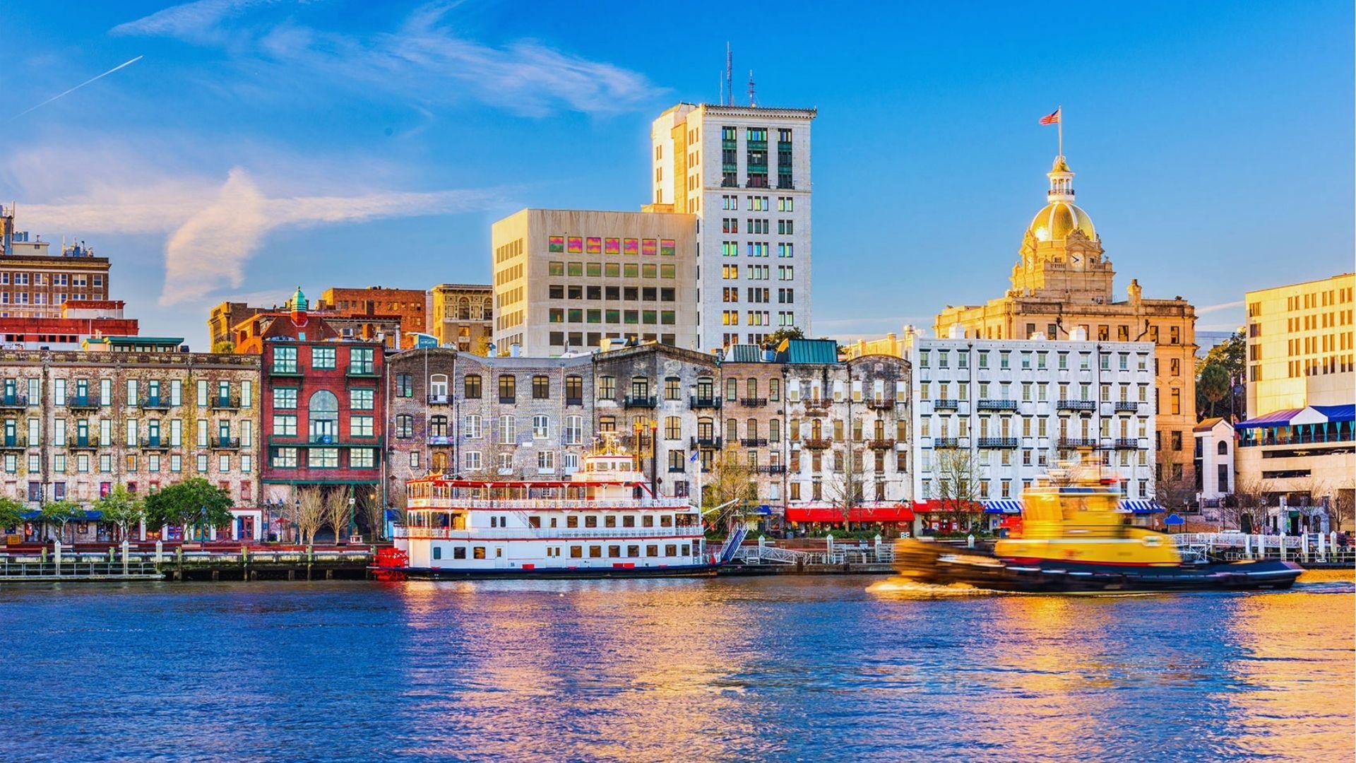 Savannah riverfront skyline with historic buildings docked riverboat moving tugboat and golden reflections on water.
